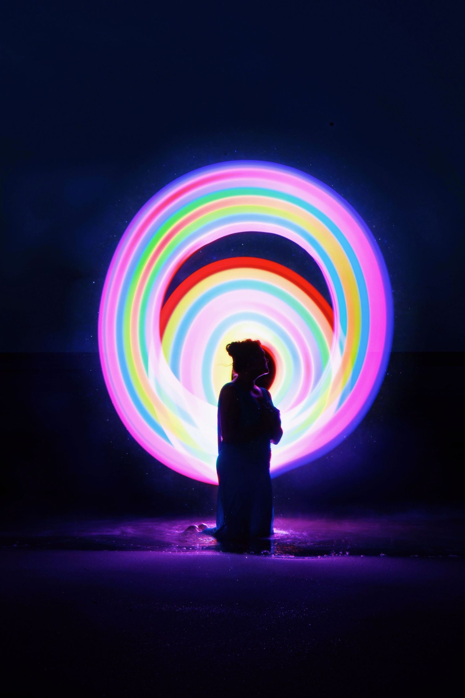 Long exposure photograph shows colorful light painting creating rainbow arcs against dark background with silhouetted figure.