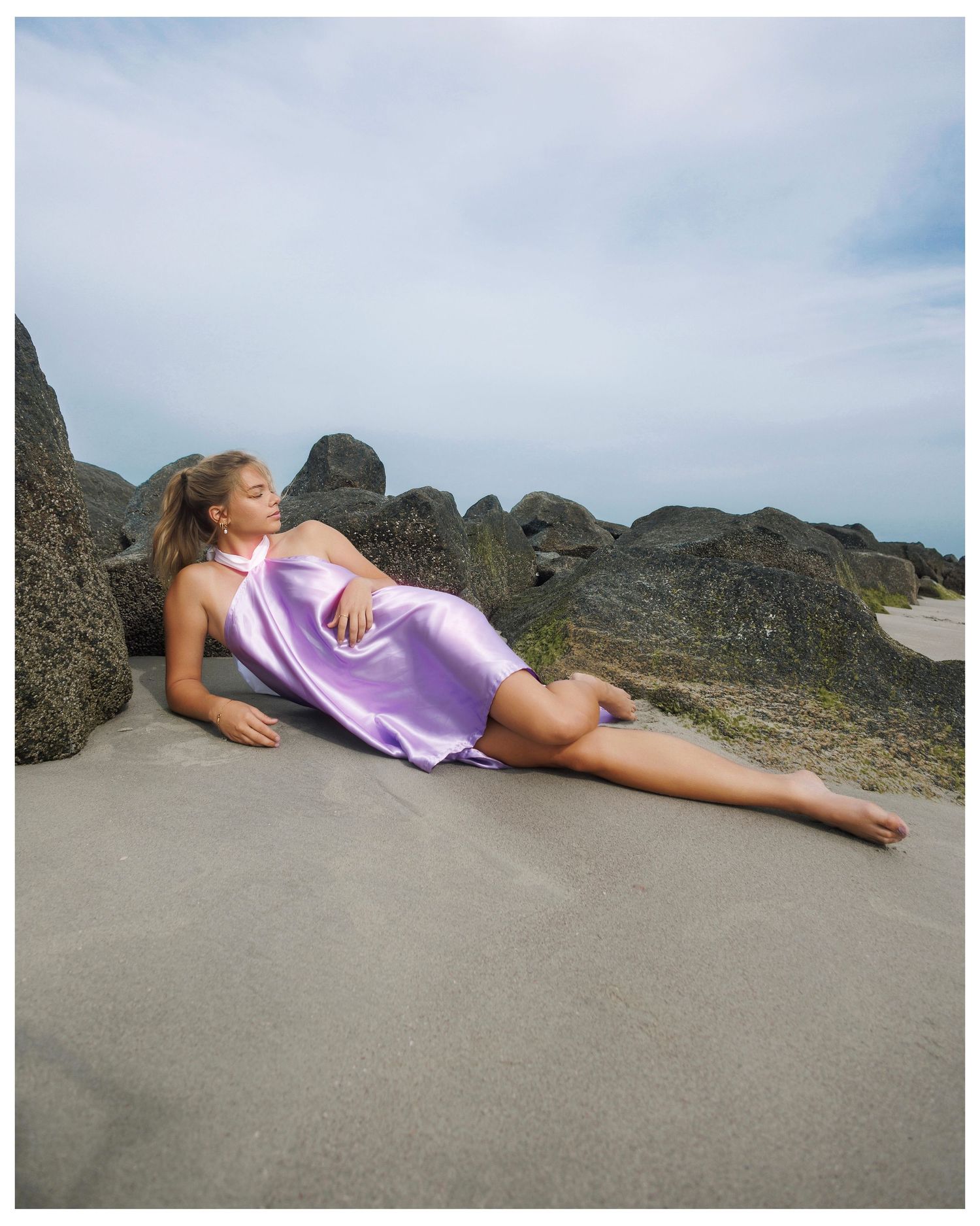 A person in a lavender dress reclines on dark coastal rocks against a cloudy sky background.