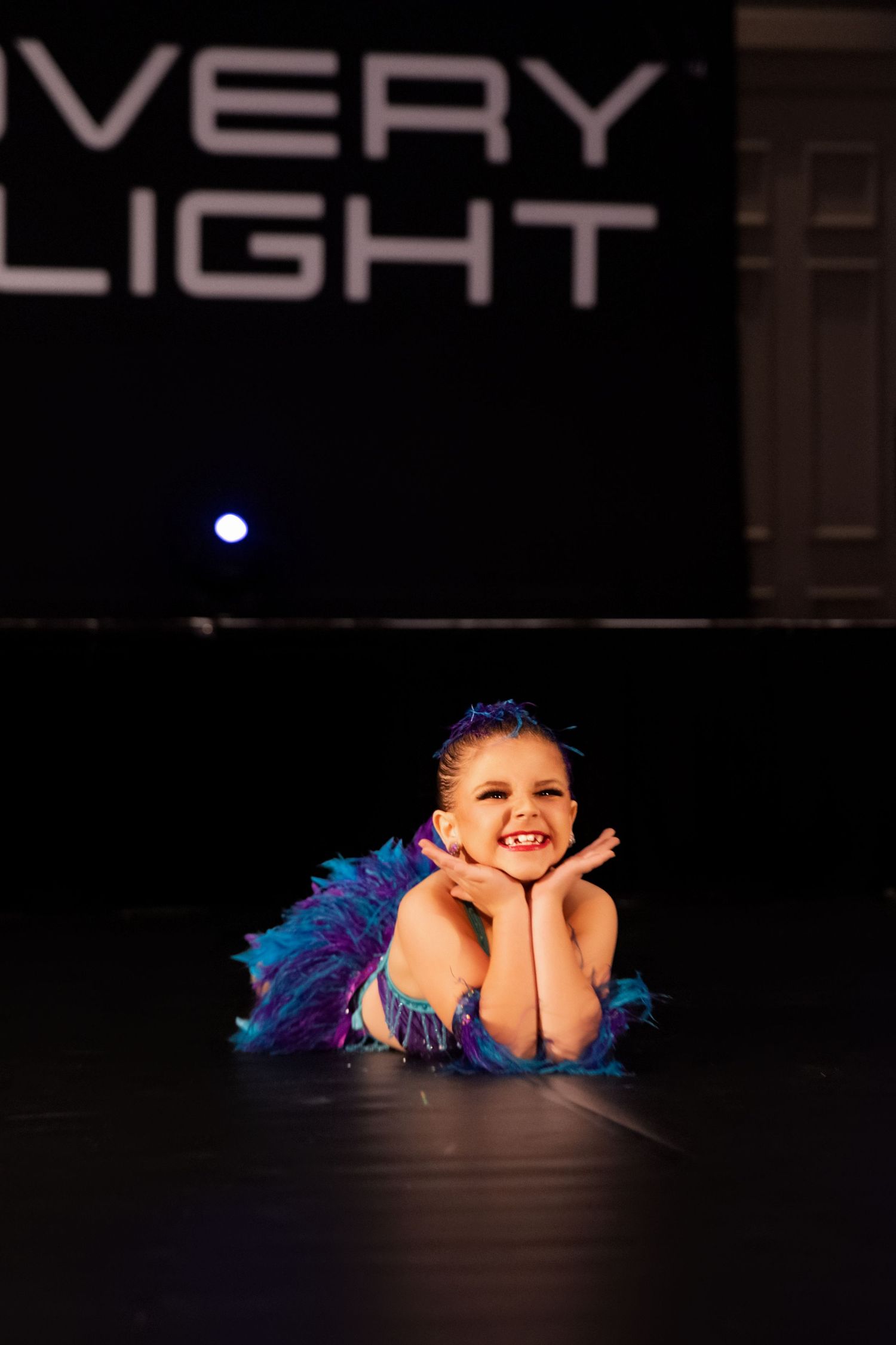 Young dancer in blue feathered costume poses on dark stage floor during performance.