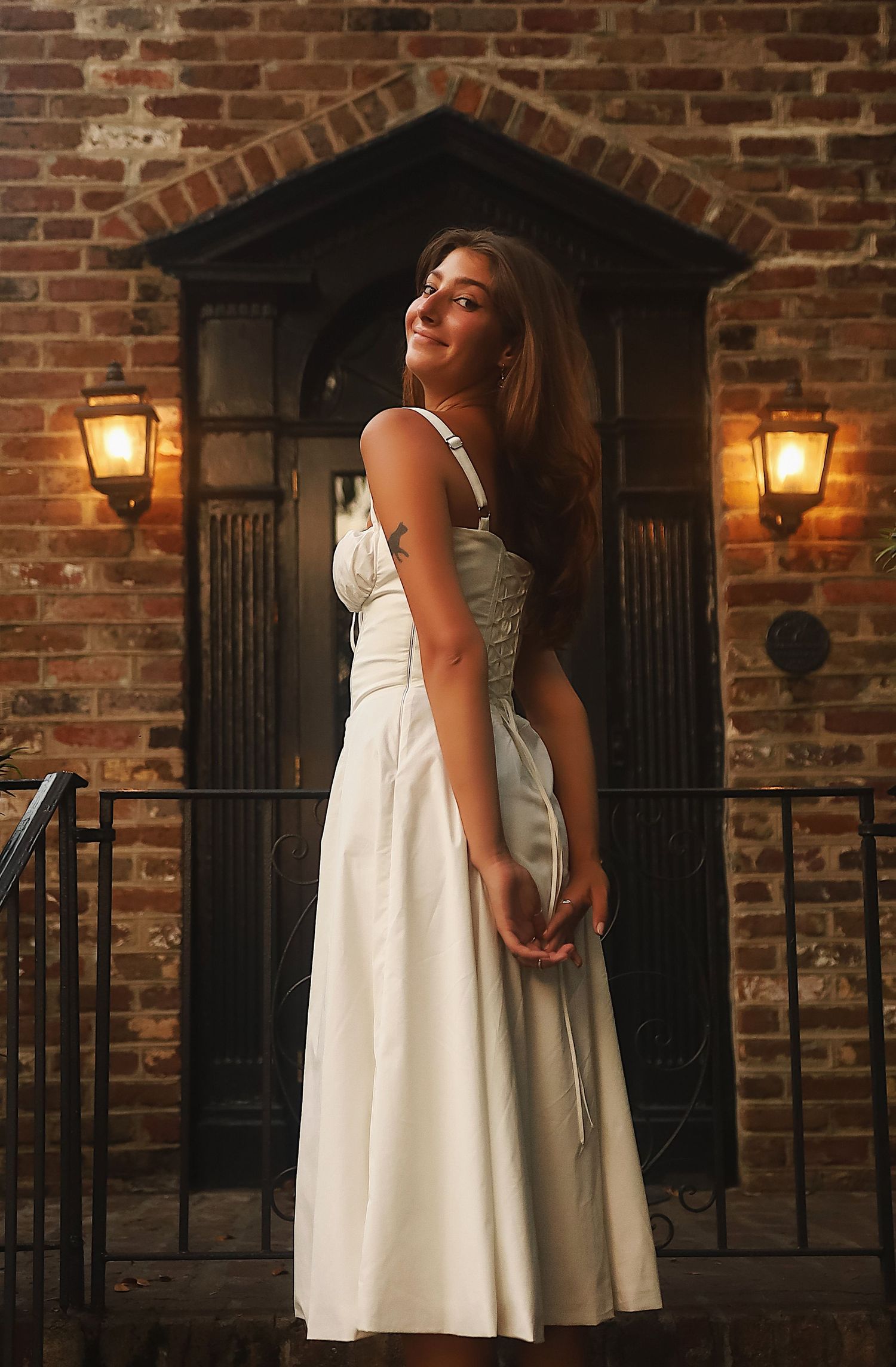 A person in a white dress stands in front of a brick wall with vintage lanterns creating a romantic evening ambiance.
