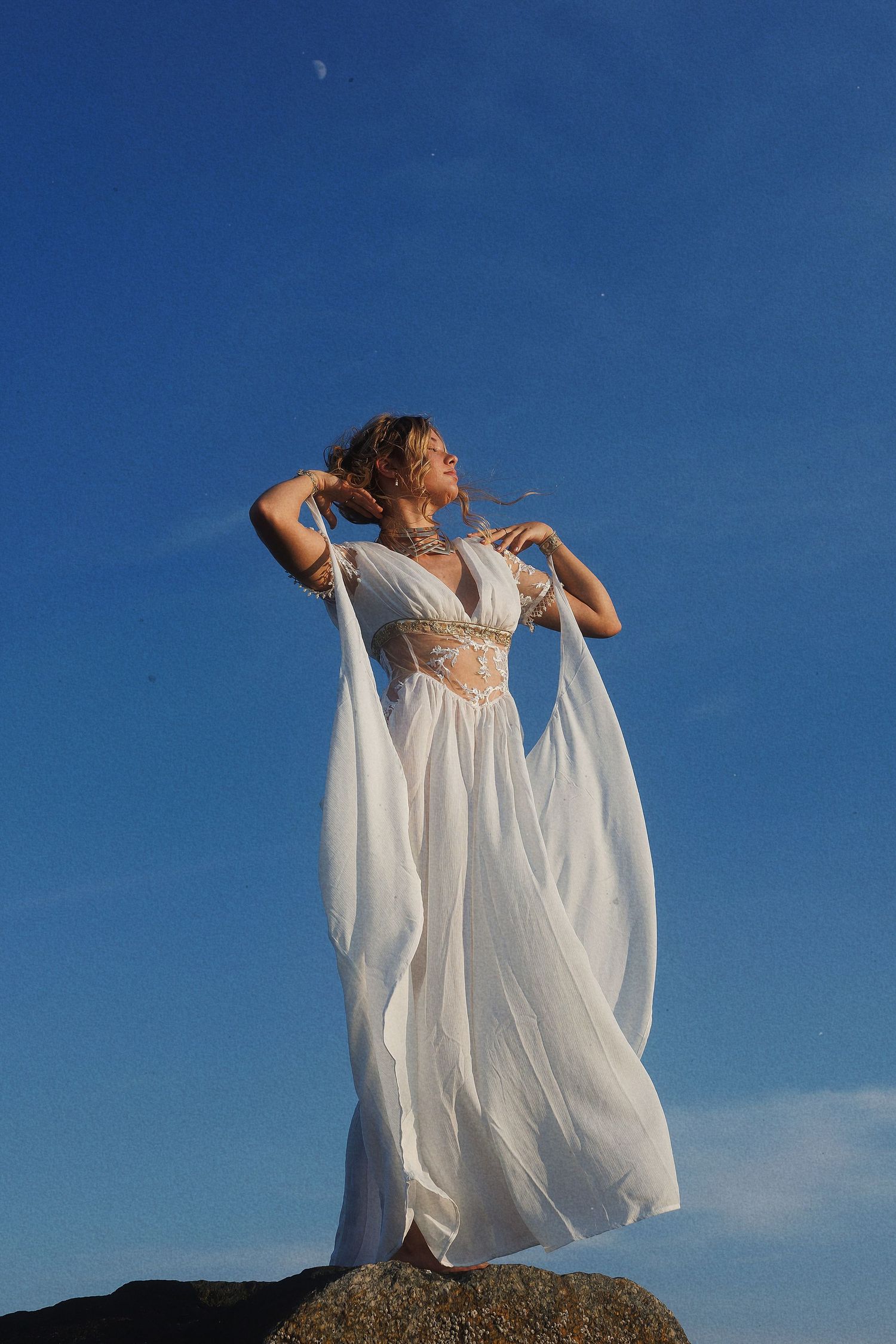Person in ethereal white dress stands gracefully atop coastal rocks against vibrant blue sky.