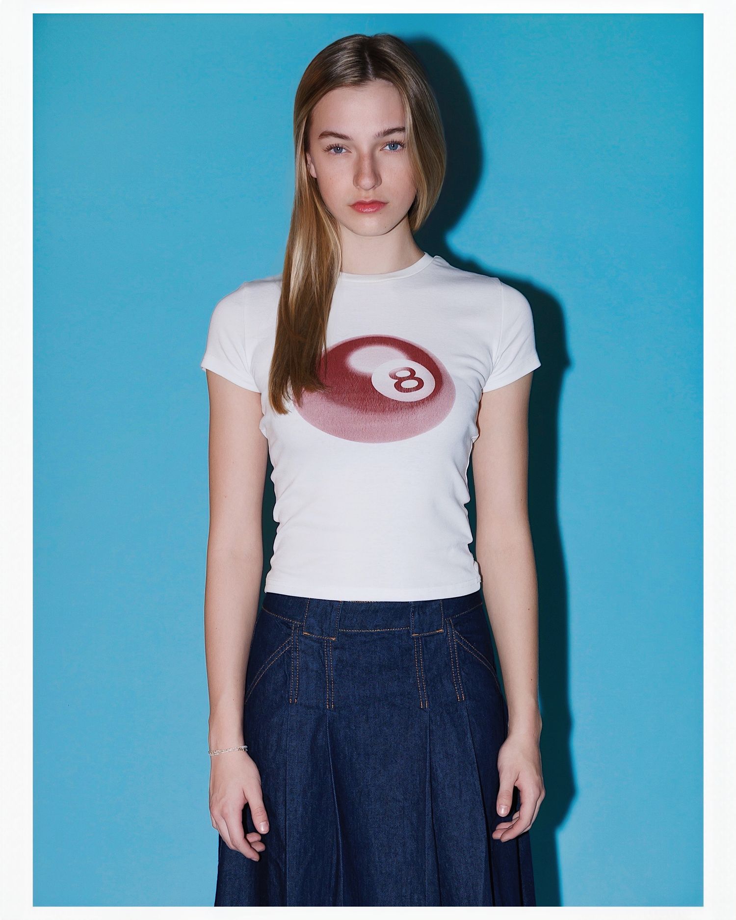 Someone wearing a white t-shirt with a printed design and denim skirt poses against a bright turquoise background.