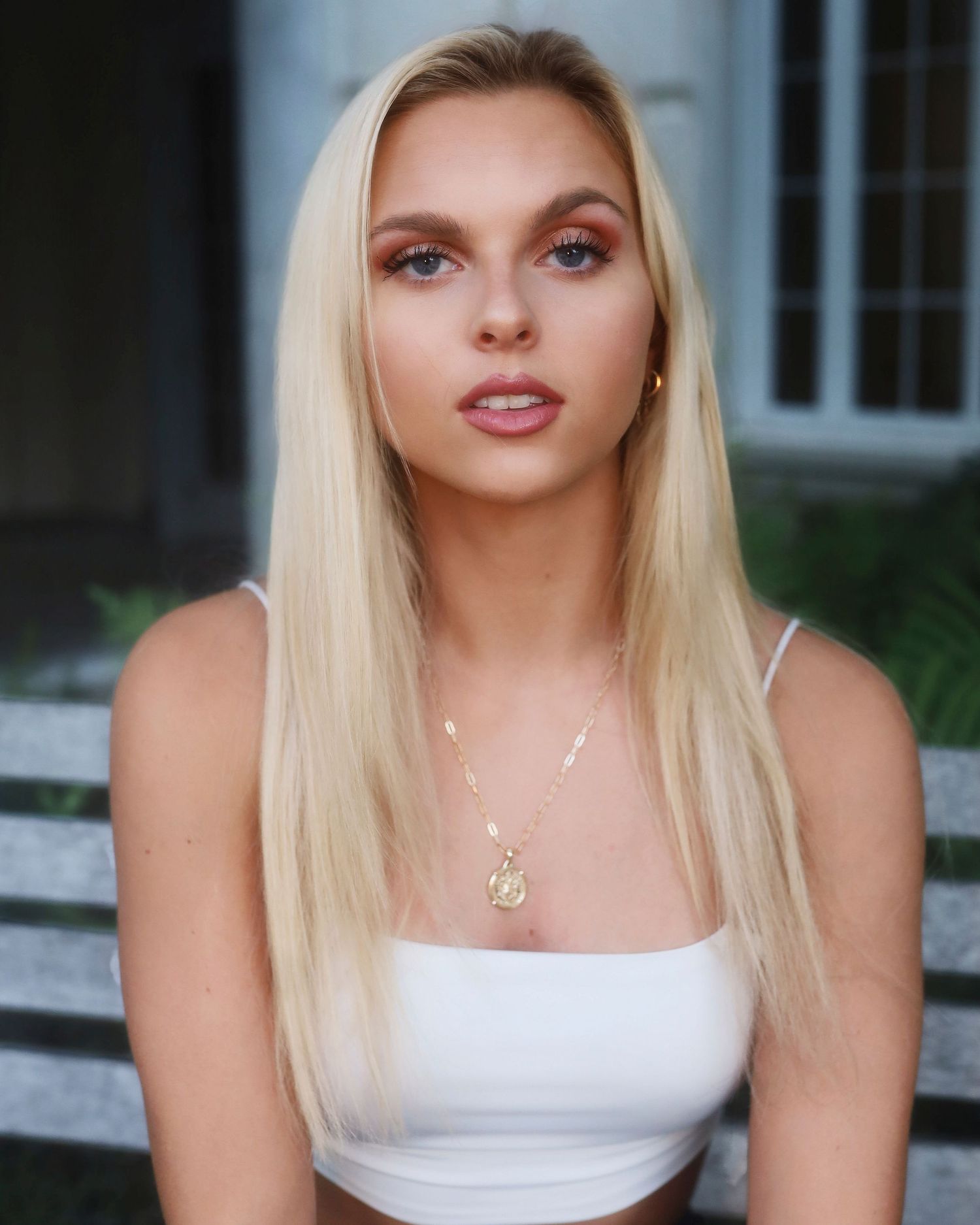 Woman with blonde hair and natural makeup wearing a white tank top and delicate necklaces.