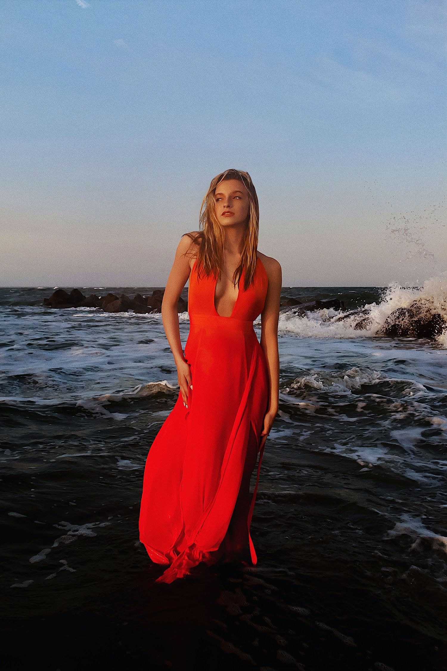 A figure in a flowing red maxi dress stands in ocean waves at sunset against a rocky shoreline.