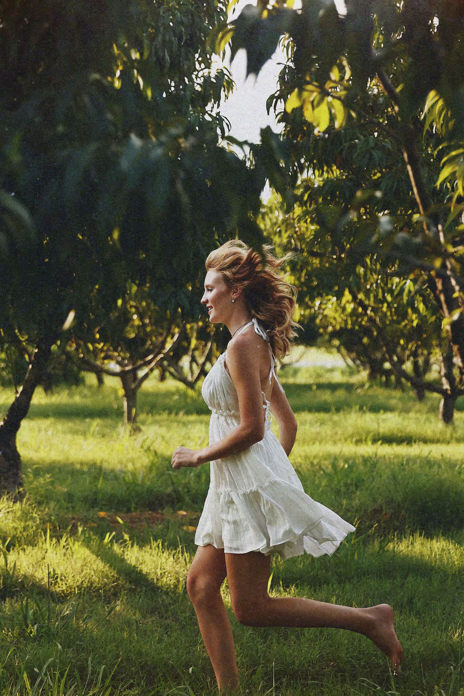 A person in a white summer dress dances and twirls through a sunlit orchard on a warm afternoon.