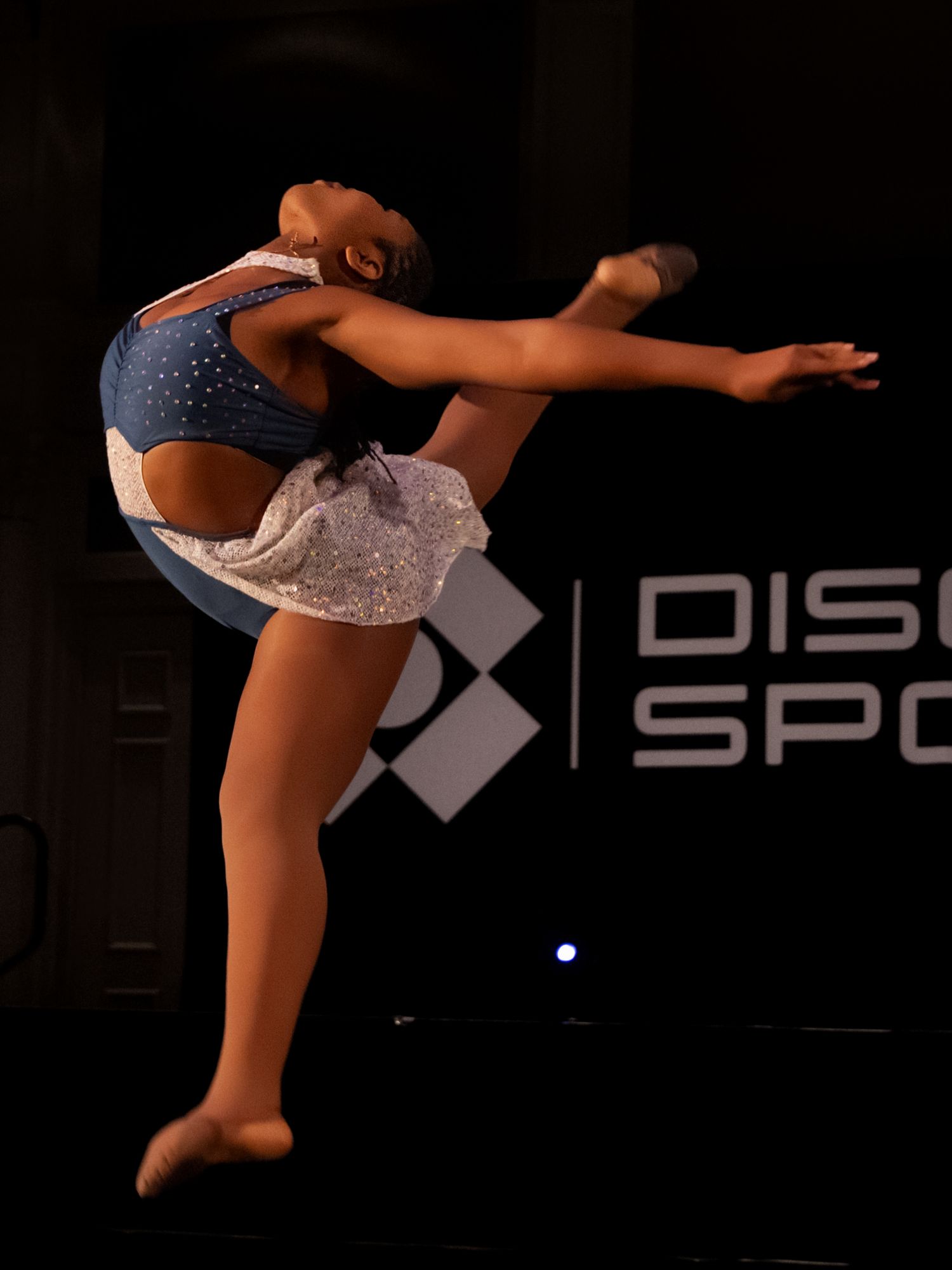 A gymnast performs a graceful aerial split leap sequence against a dark background with illuminated signage.