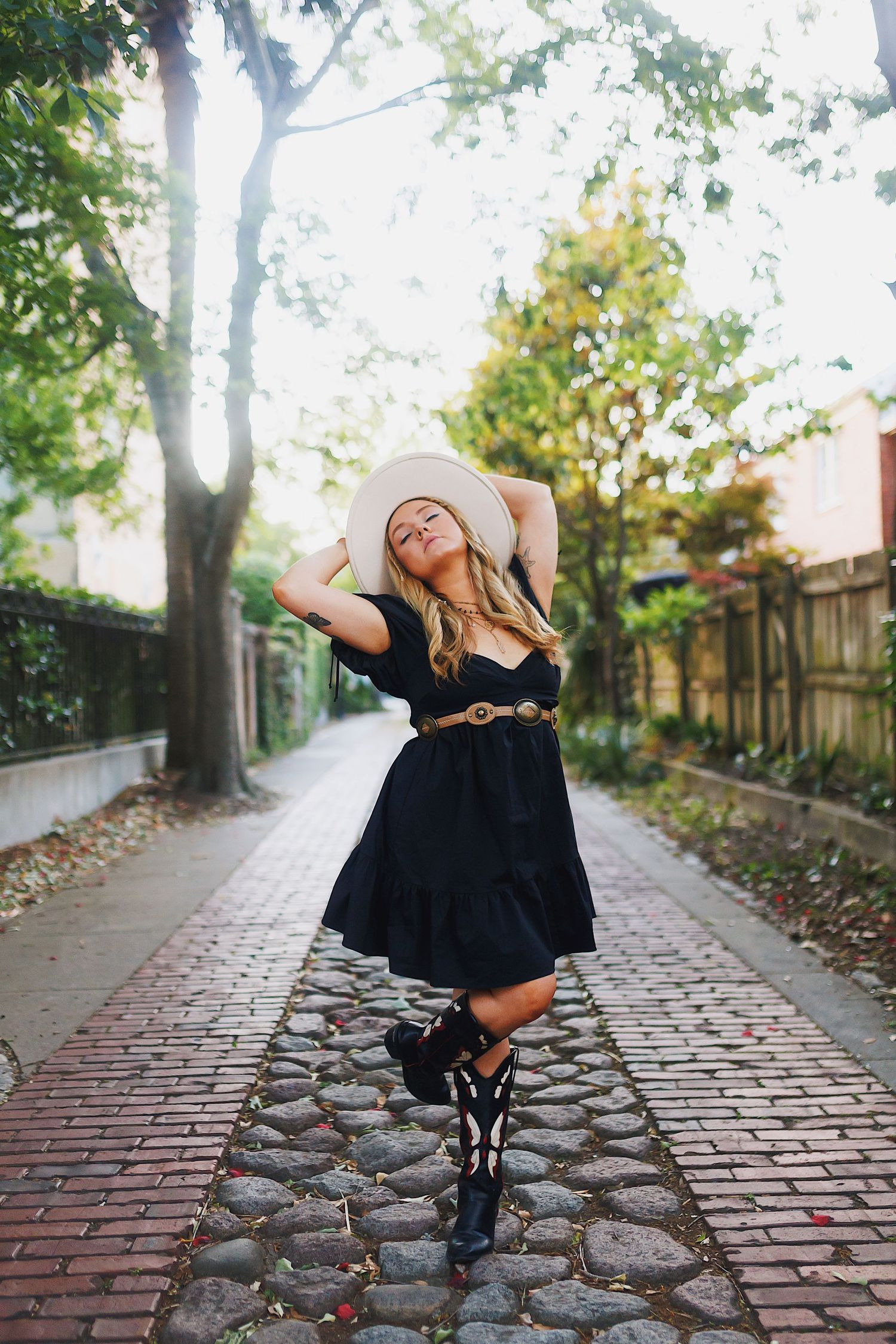 A person in a black dress poses creatively on a cobblestone path surrounded by trees and autumn leaves.