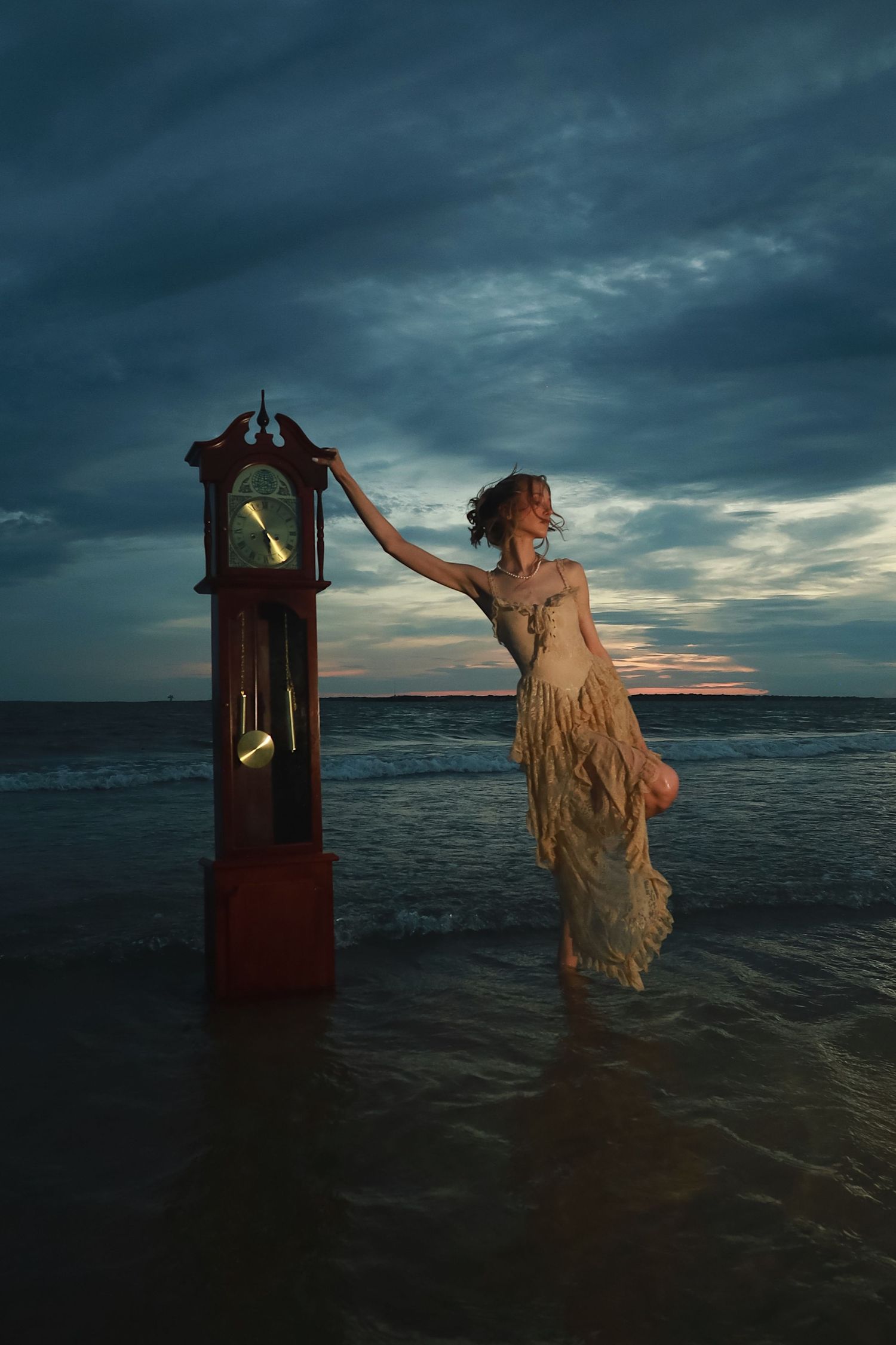 A figure in a flowing dress stands in ocean water beside a red wooden phone booth at sunset.