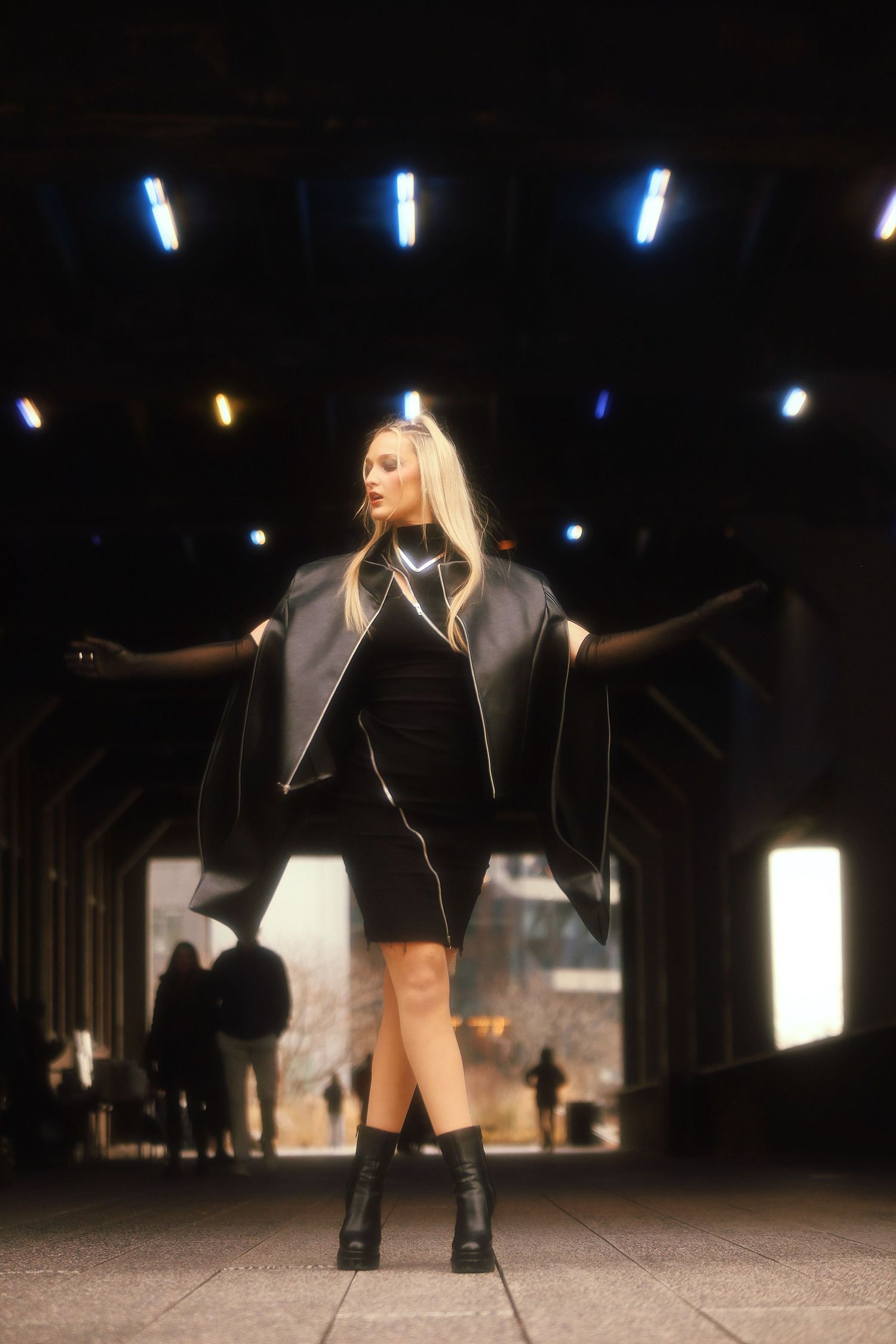 A performer in a short black dress and leather jacket strikes a dramatic pose under stage lighting.