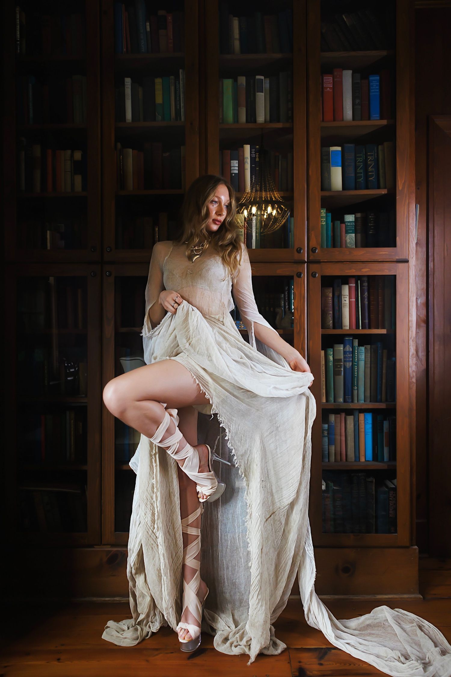 Artistic boudoir photoshoot in front of vintage wooden library bookshelves features flowing white fabric and candlelight.