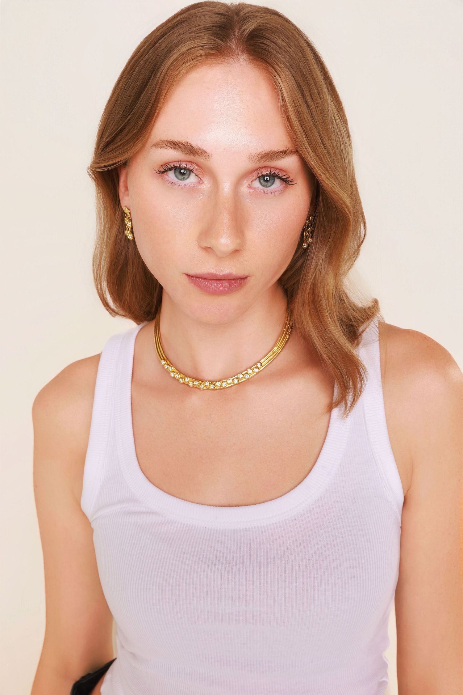 A person wearing a white tank top accessorized with a gold chain necklace and earrings against a light background.