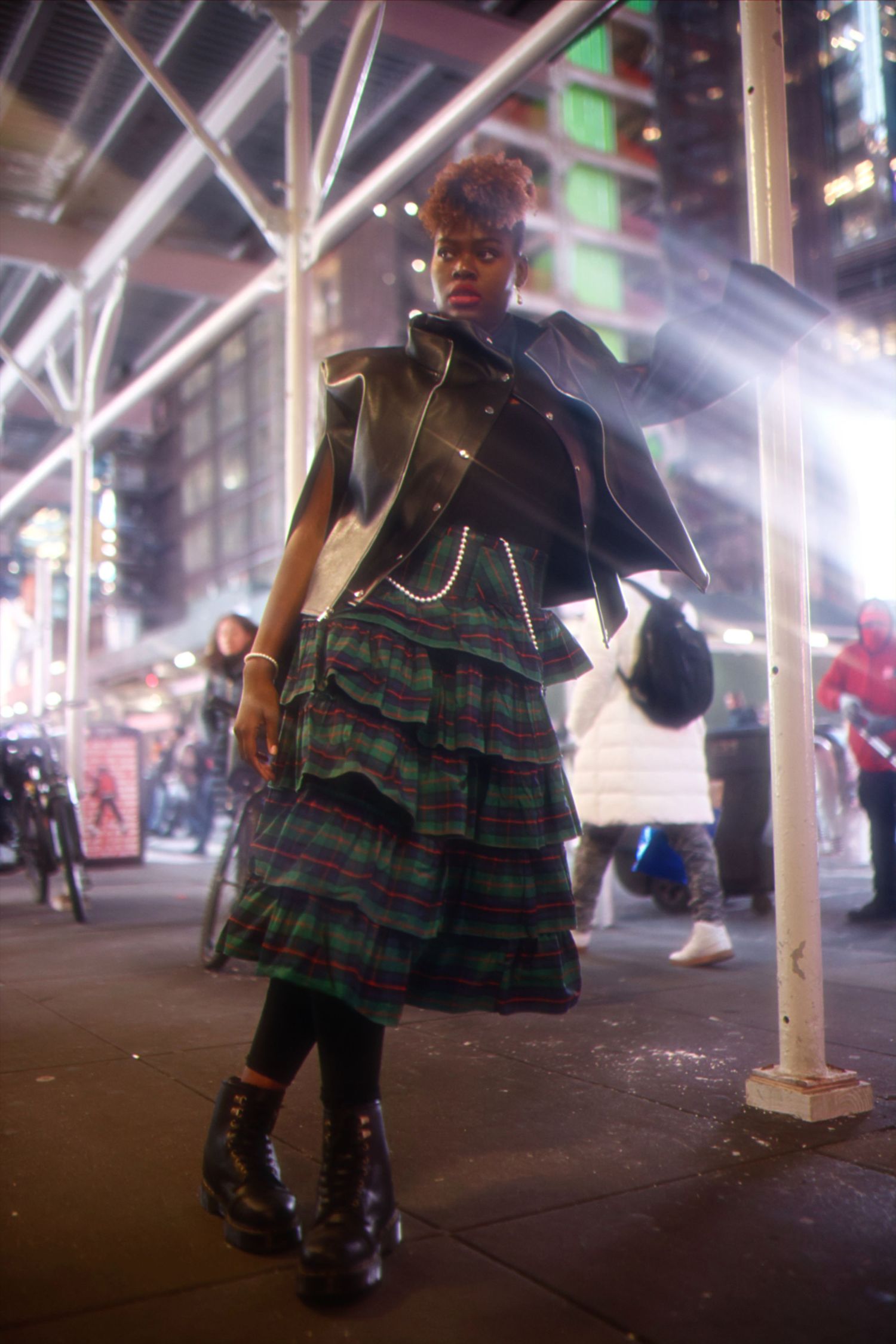 Fashion portrait at night showing plaid pleated skirt, black jacket and combat boots on city street corner with neon lights.