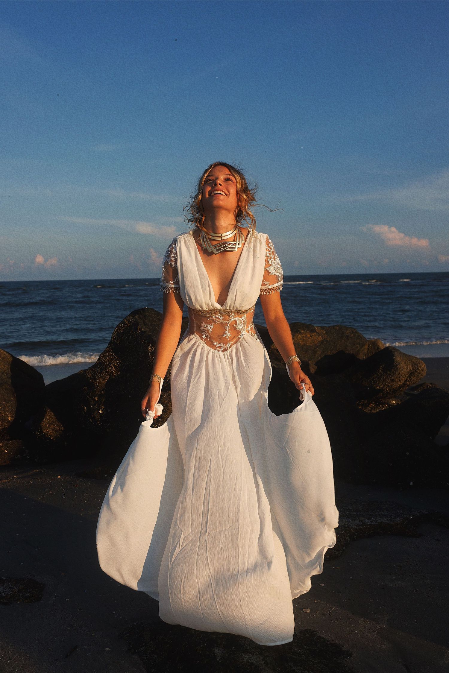 A person in a flowing white dress poses against ocean rocks at sunset, creating a dreamy beachside scene.
