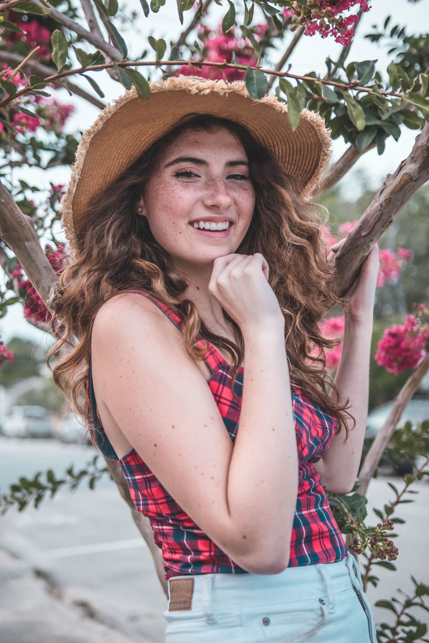 Someone in a plaid top and straw hat poses playfully near flowering trees in a casual outdoor photoshoot.