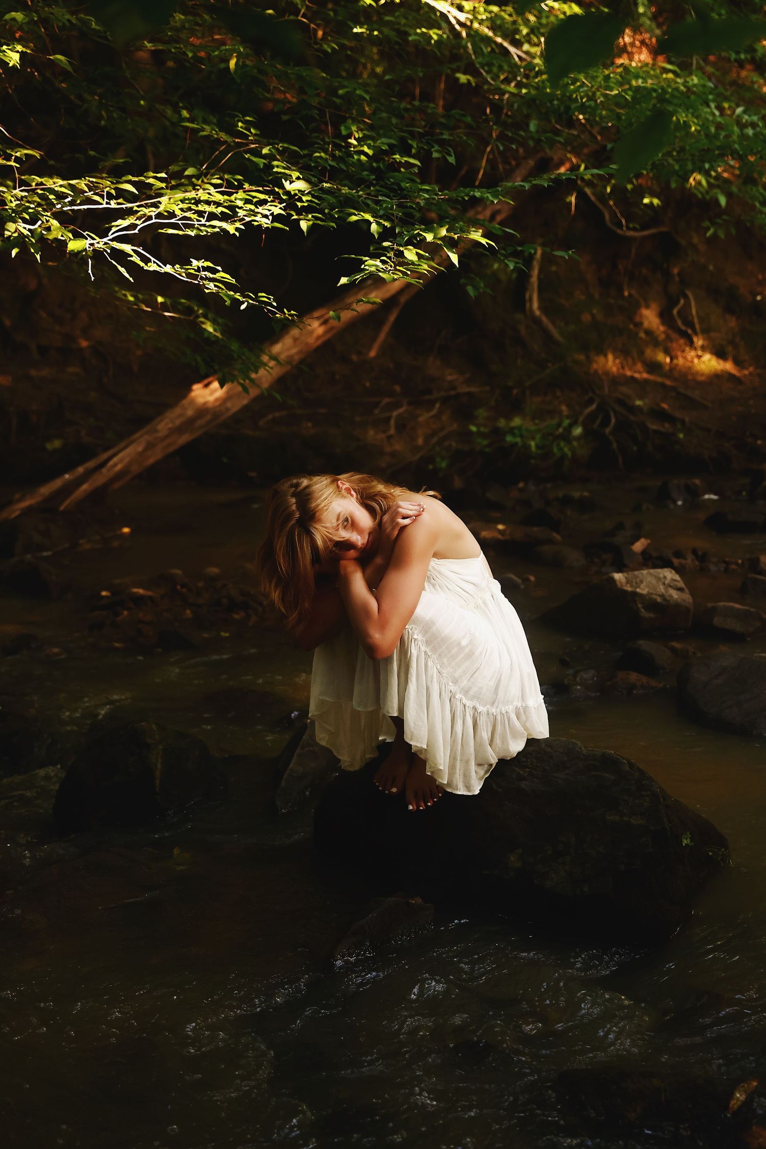 A figure in a white dress sits perched on dark rocks near a stream in a moody forest setting.