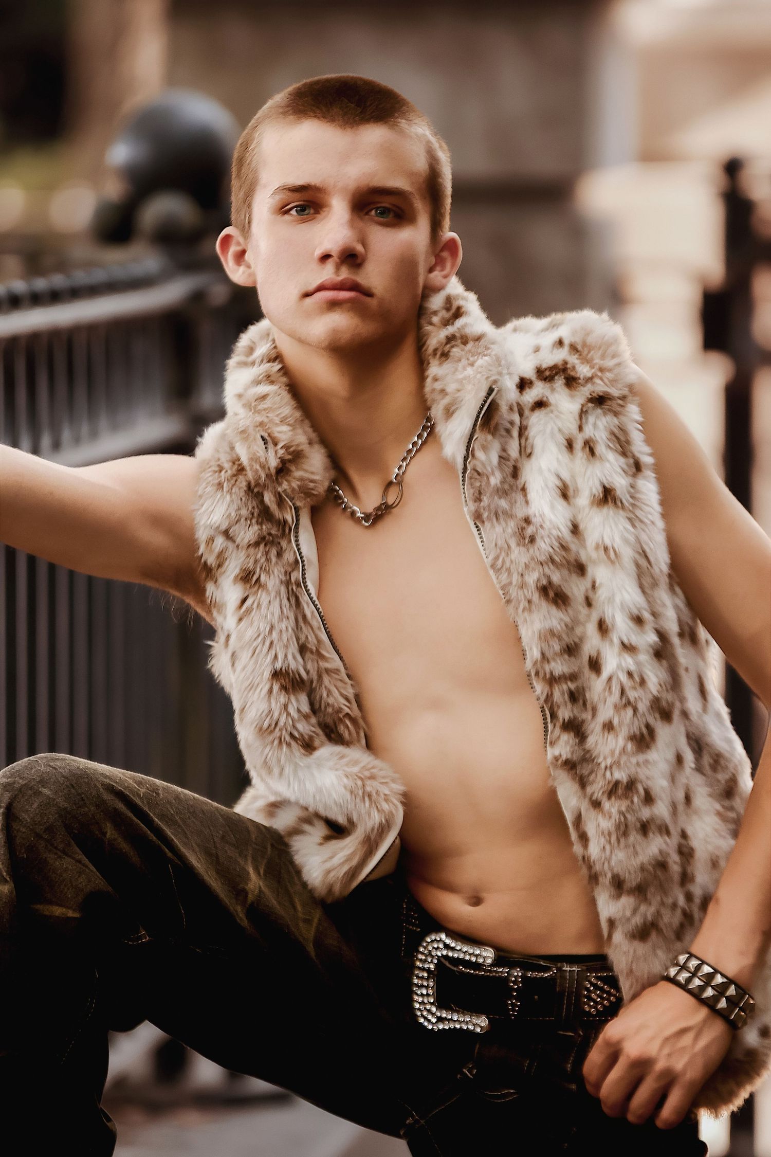 Young model poses outdoors wearing a spotted fur vest and dark pants with a decorative belt buckle.