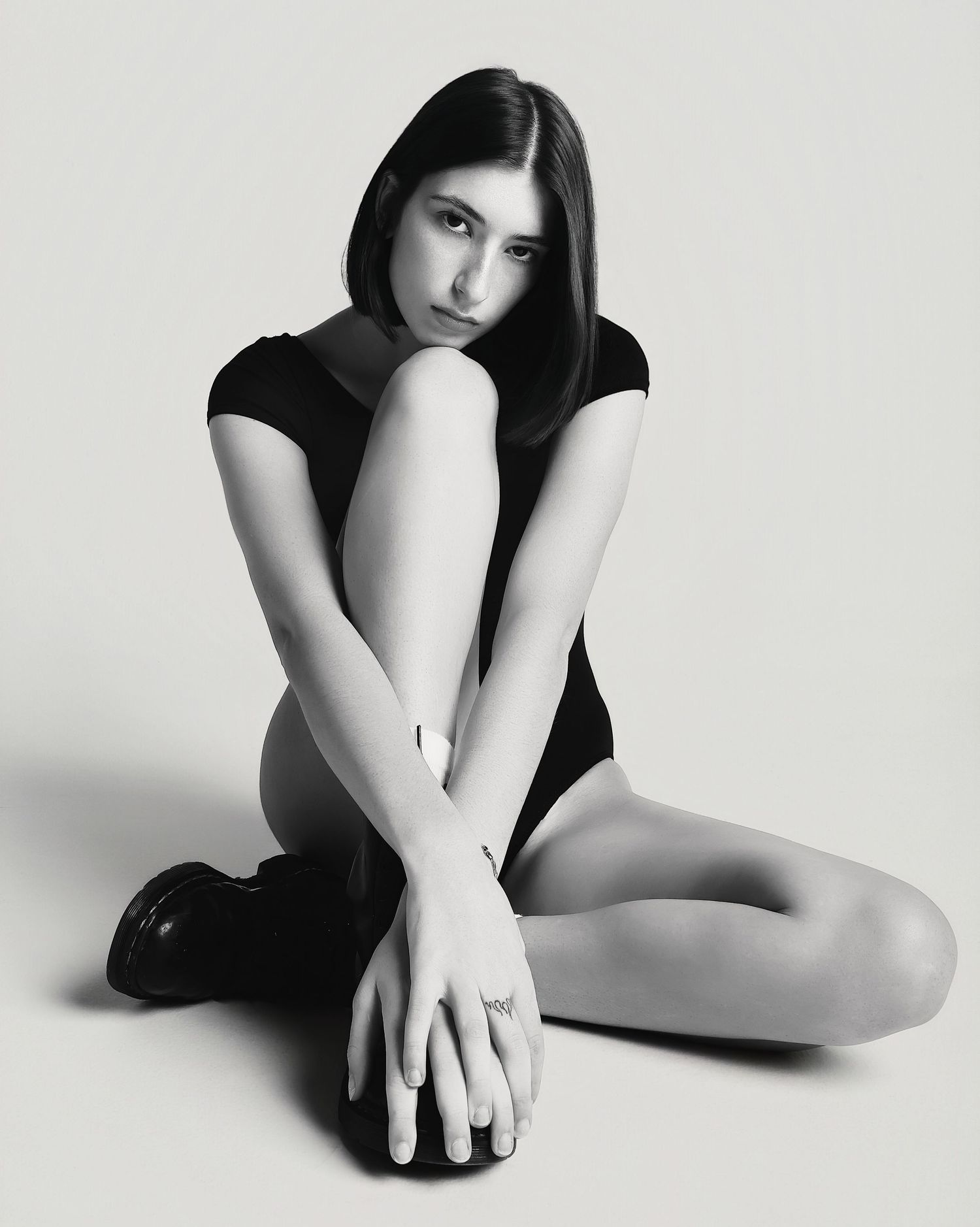 Black and white artistic portrait of a person in a dark outfit sitting on the floor in a minimalist pose.