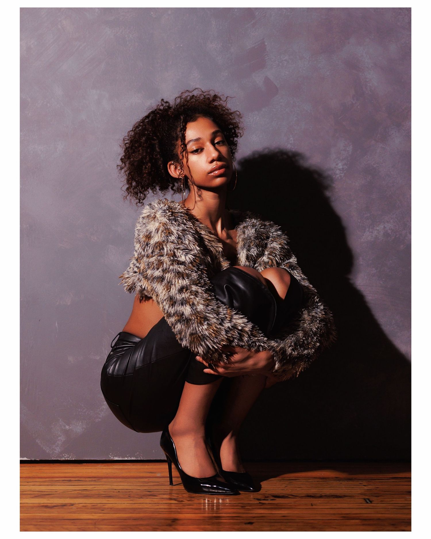 Fashion portrait with model in textured fur jacket and black heels crouching on wooden floor against gray backdrop.