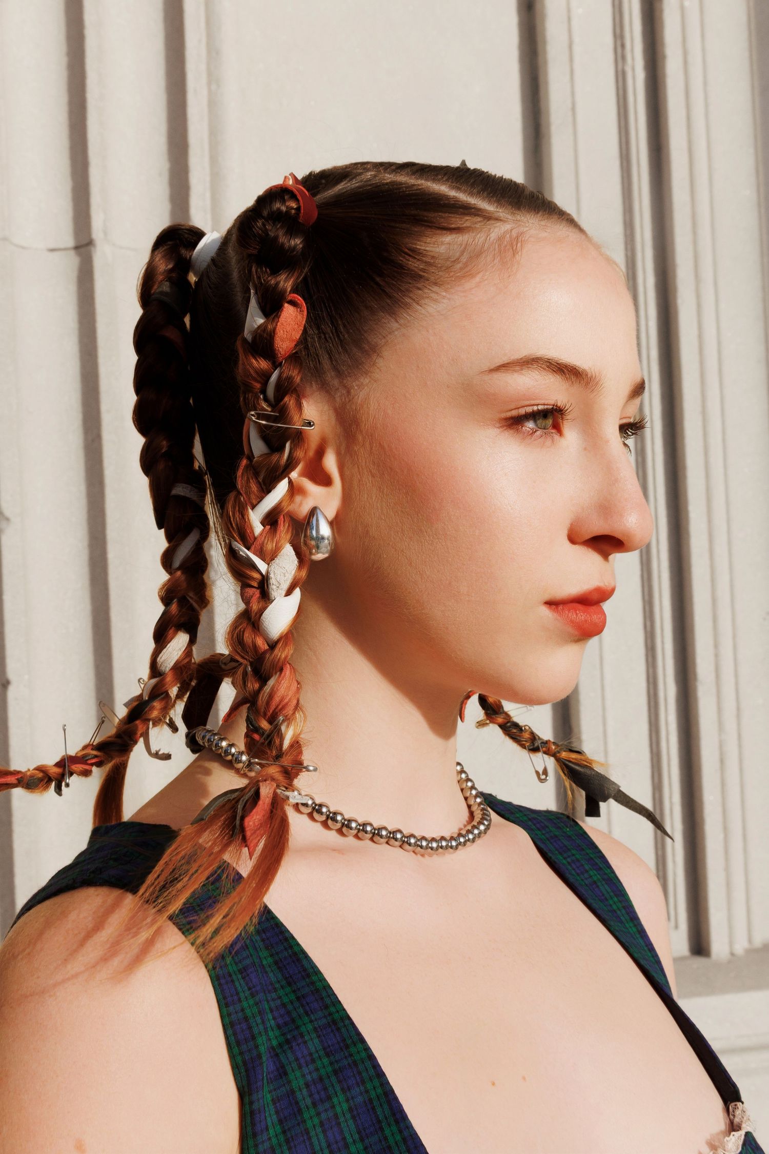 Side profile portrait with braided hairstyle adorned with ribbons, wearing plaid top and chain necklace in warm lighting.