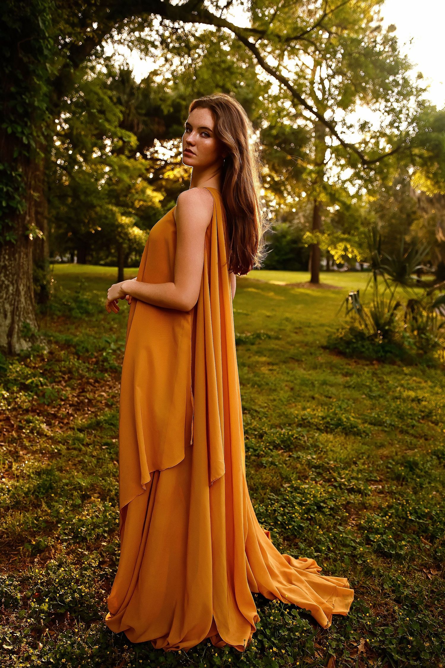 A person wearing a flowing mustard yellow dress poses in a garden at golden hour with sunlit trees in the background.