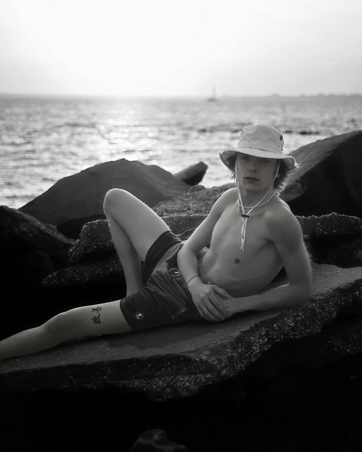 A shirtless person in a sun hat relaxes on dark rocks by the ocean in black and white photography.