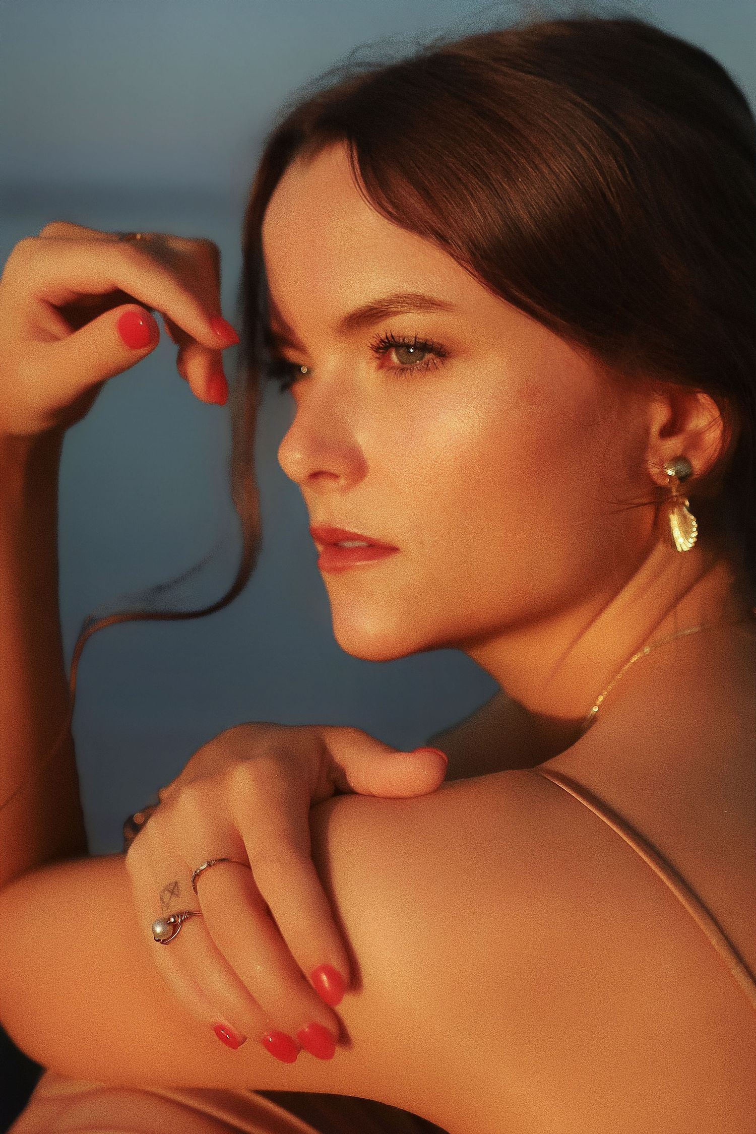 Profile view portrait with warm lighting shows gold earrings, red lipstick and nails against a dusky blue background.