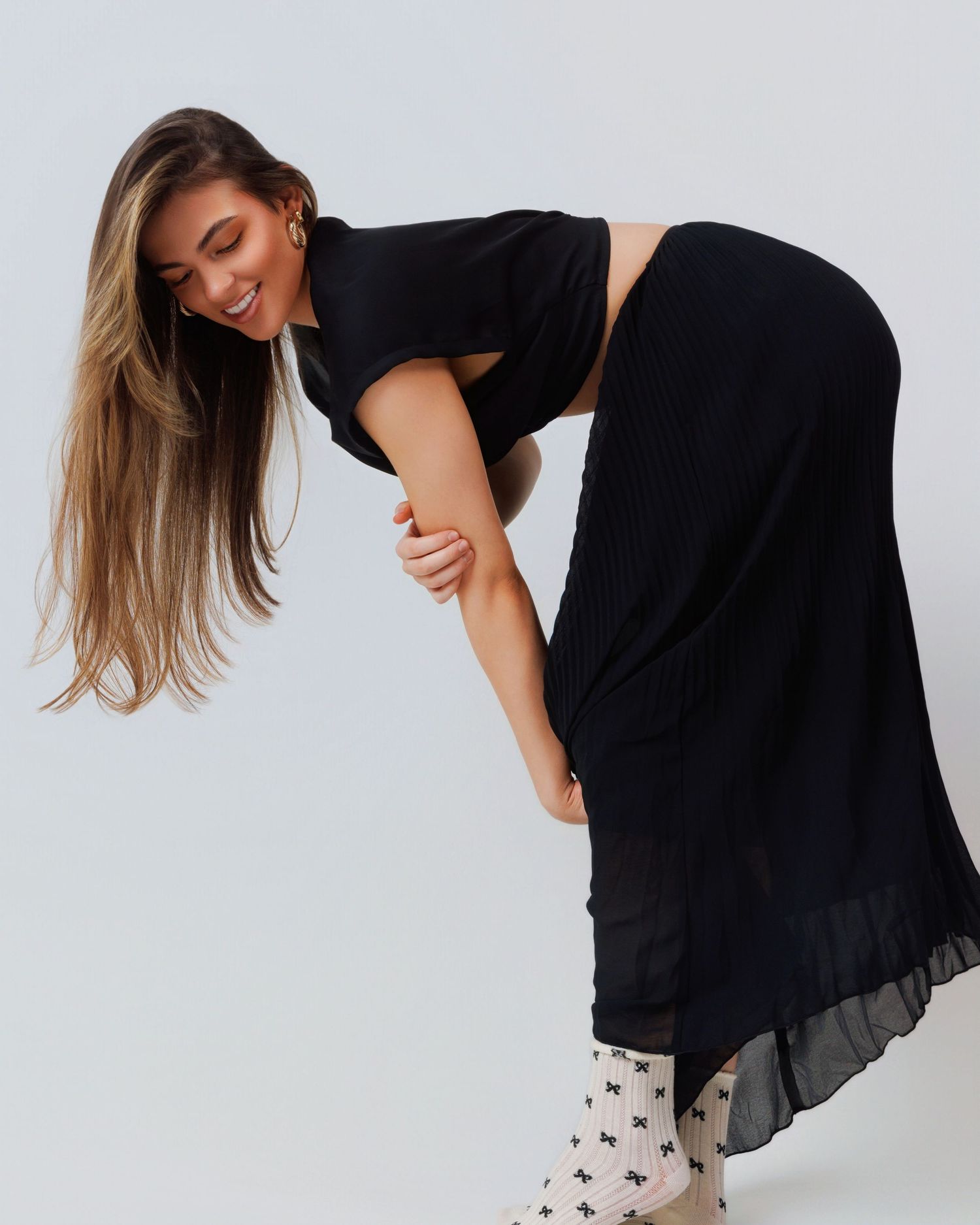 A model in a black dress and patterned socks leans forward playfully against a white backdrop.