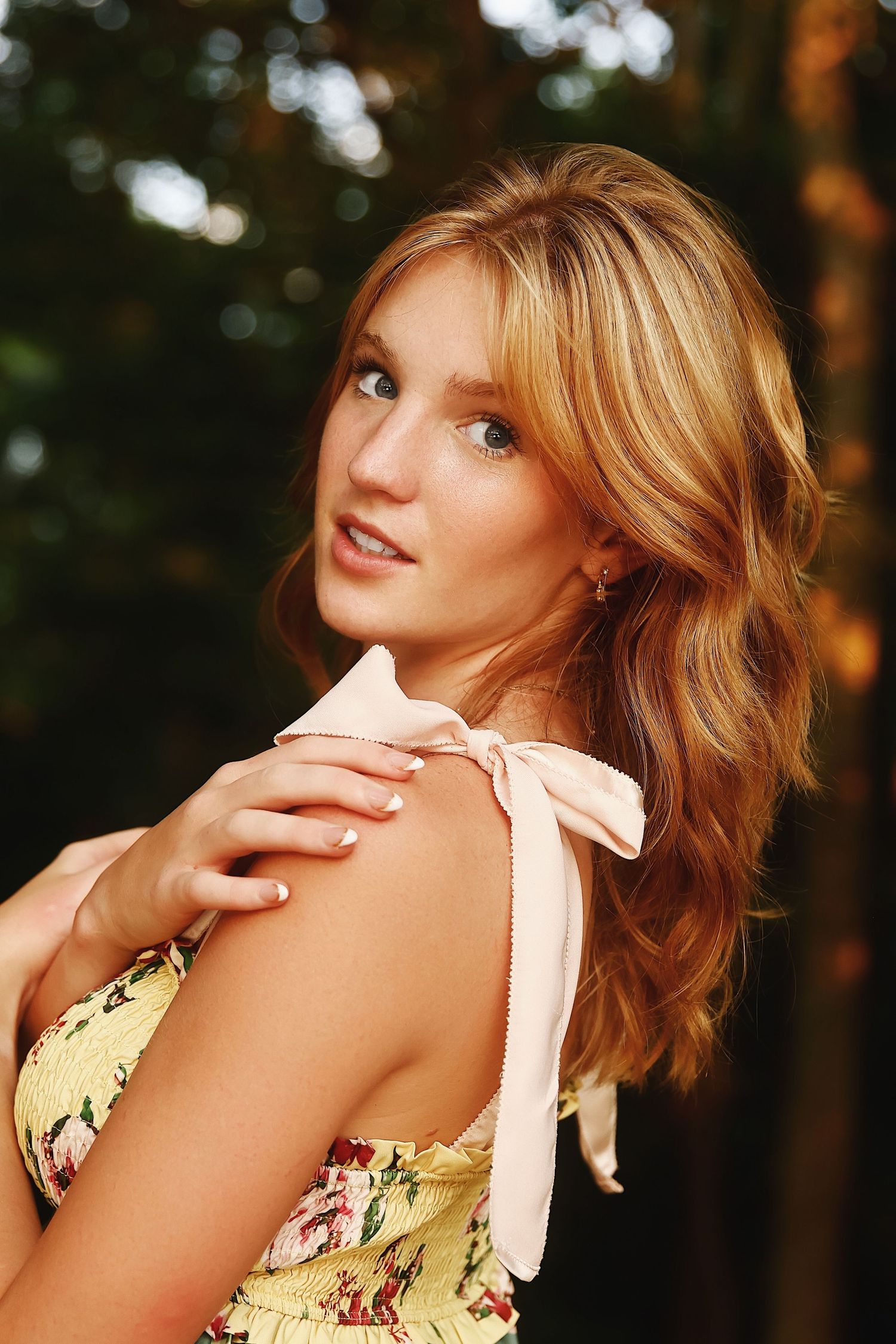 A person in a yellow floral dress poses playfully in a forest setting during golden hour lighting.