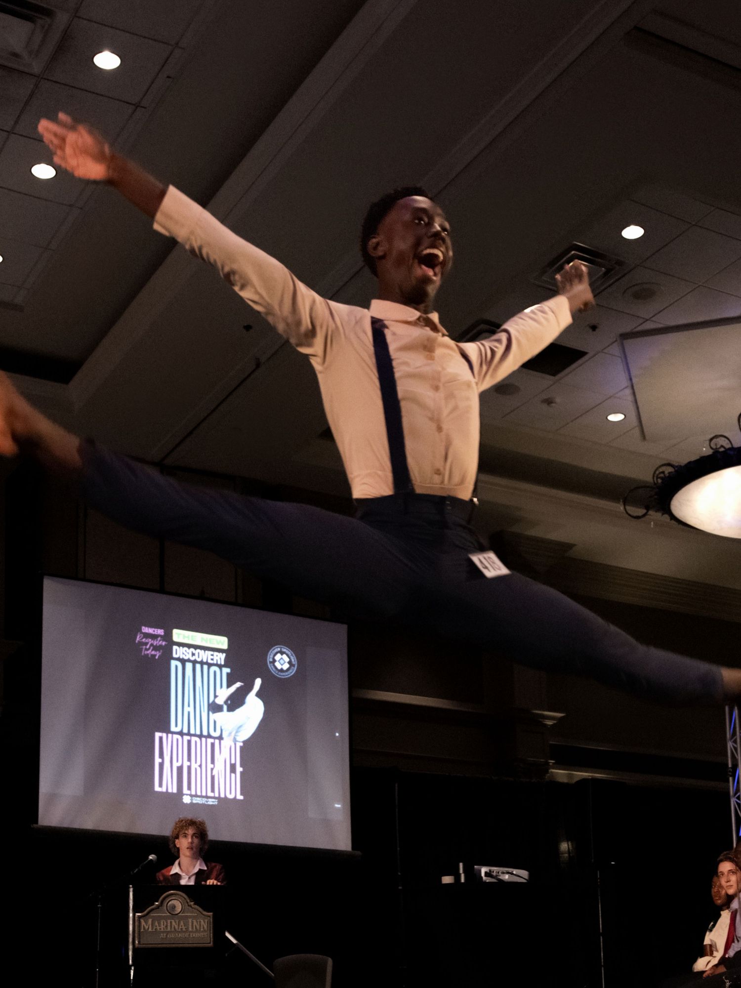 Speaker gestures enthusiastically on stage with arms raised while presenting in front of projected screen display.