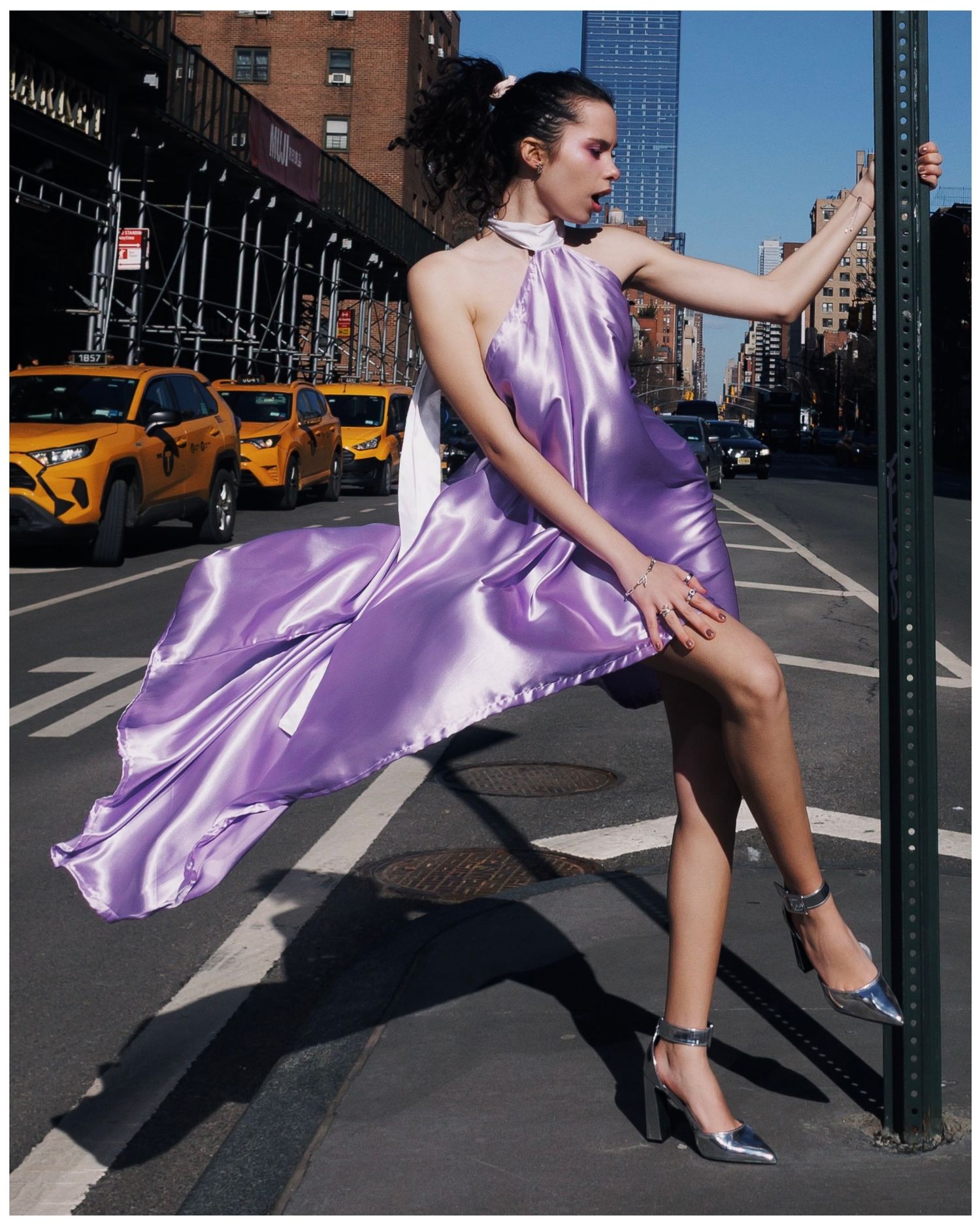 A flowing purple satin dress billows dramatically across a New York City street with yellow taxis in the background.