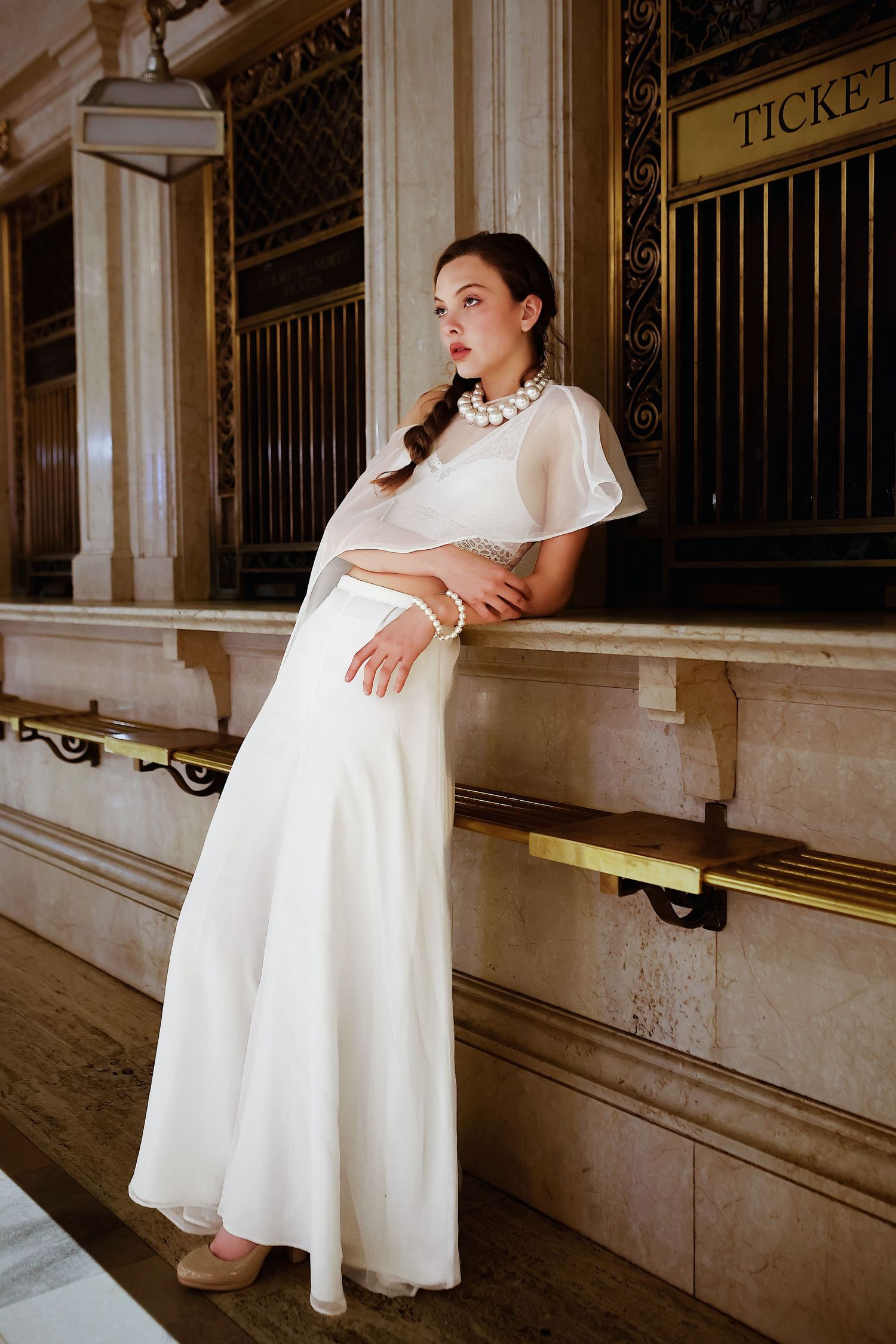 Fashion model in vintage-inspired floor-length white dress and statement necklace poses elegantly on marble steps.