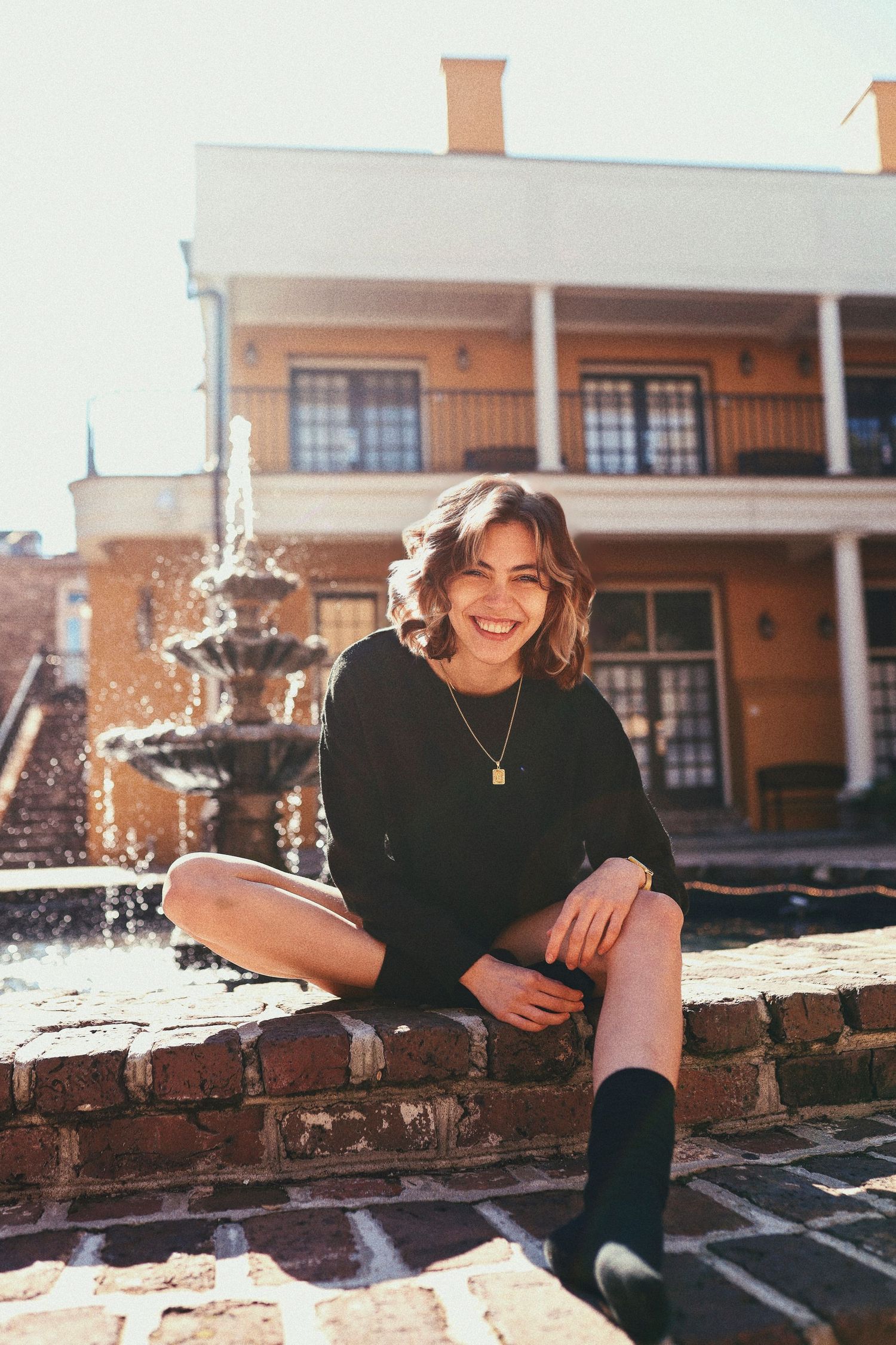 A person in a black sweater sits smiling on a brick ledge near a fountain with a colonial style building in the background.