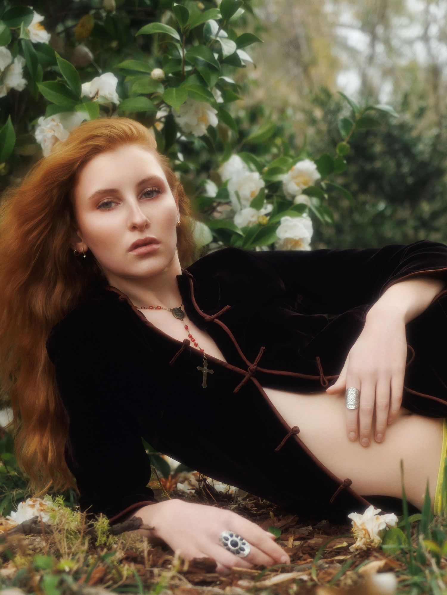 A person in black velvet clothing reclines on the ground near blooming white flowers and green foliage in a dreamy outdoor scene.