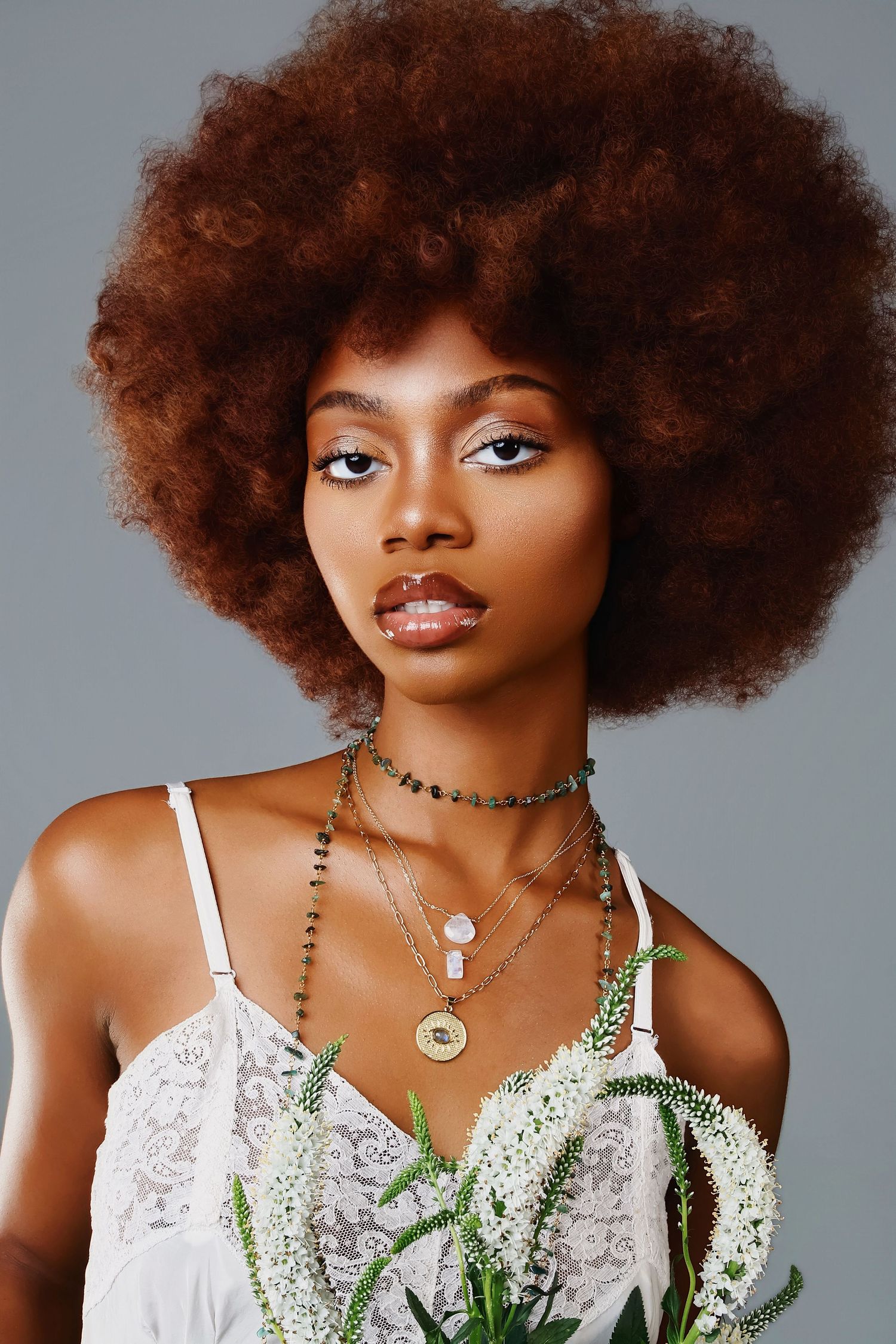 Studio portrait showing a natural afro hairstyle against grey background with delicate white lace top and layered necklaces.