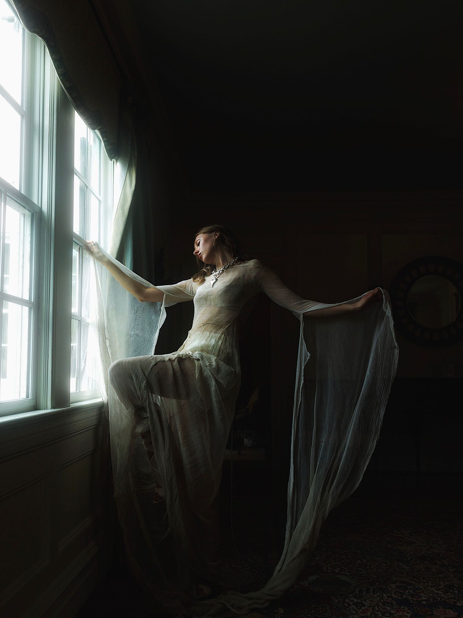 Figure in a flowing white dress poses gracefully by a window in dramatic light and shadow.