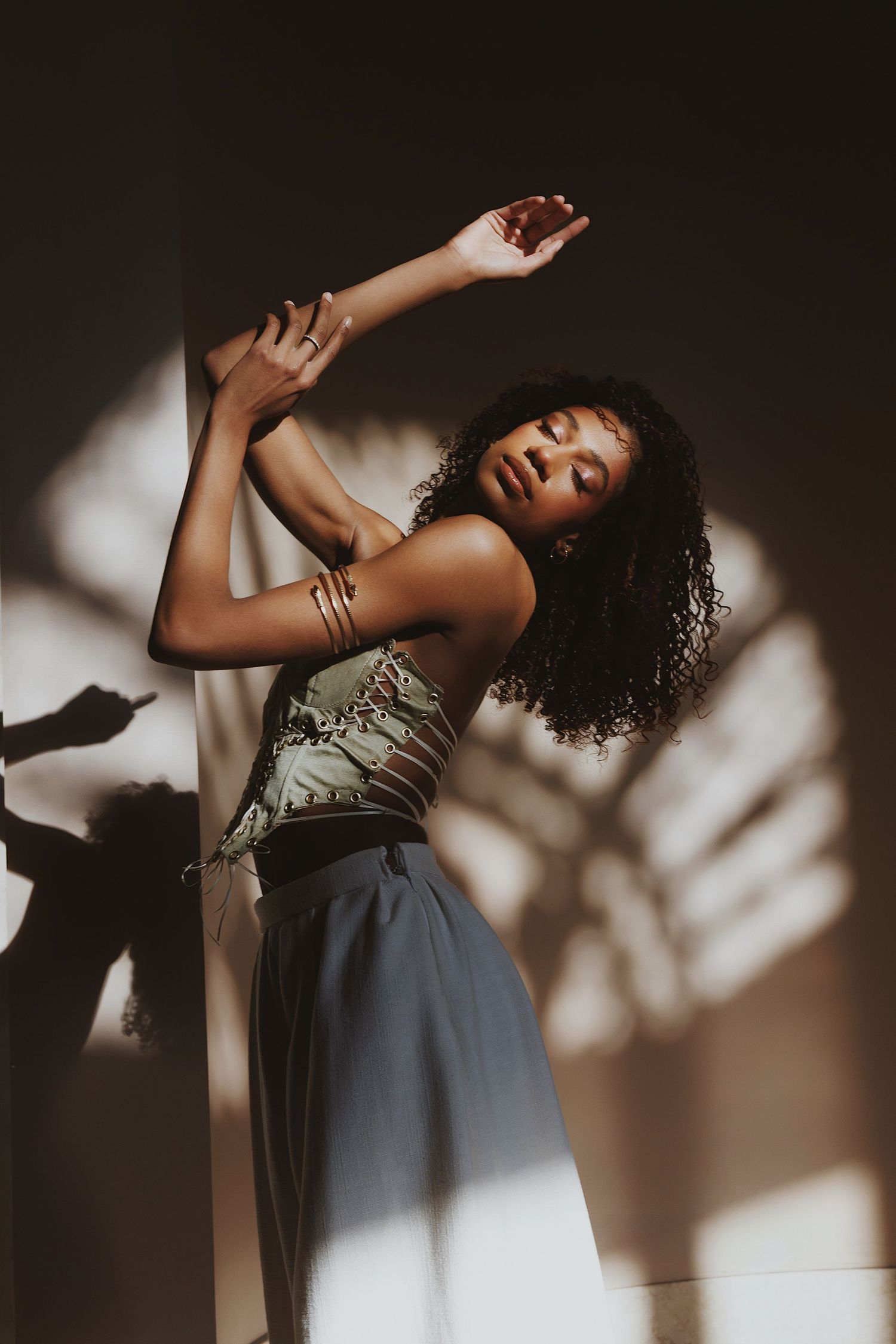 A person in a green top and gray skirt poses dramatically against a wall with shadow patterns in moody lighting.