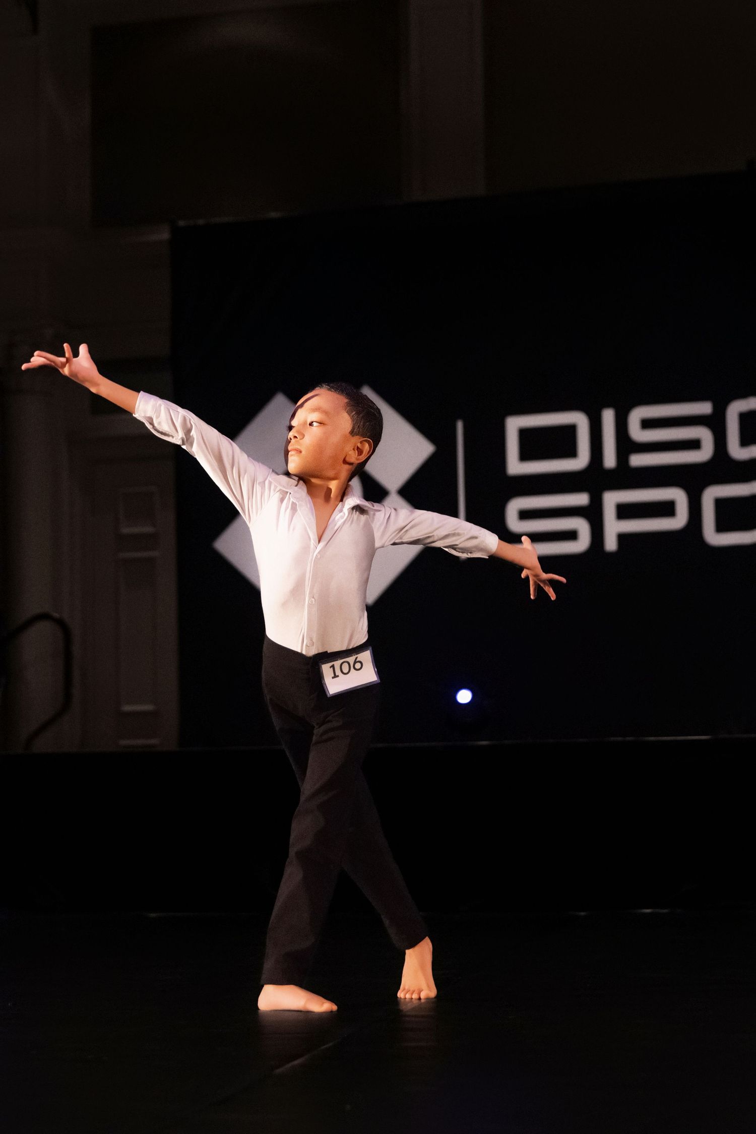 A young dancer in black pants and white shirt performs an expressive contemporary dance pose on a dark stage.