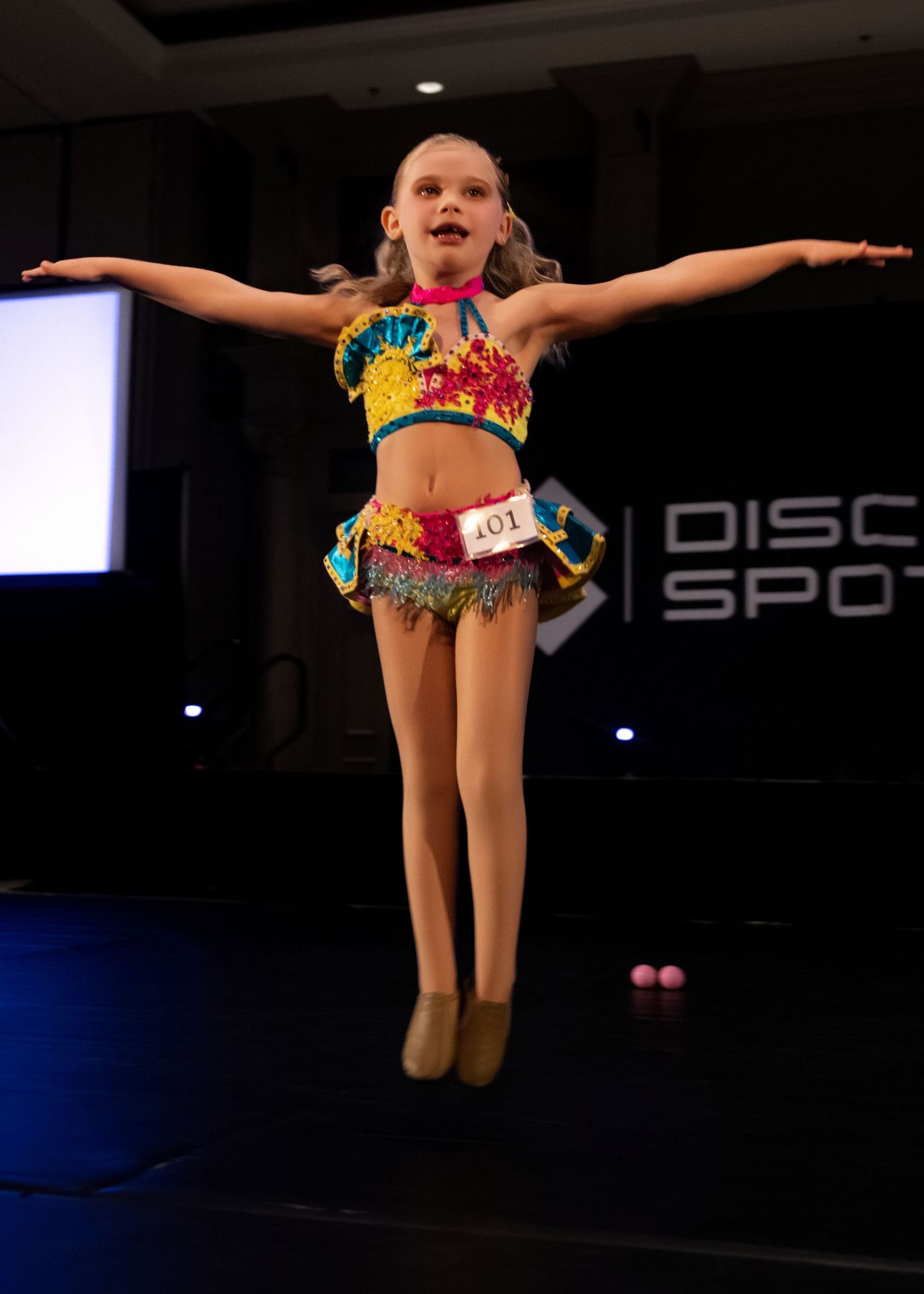 Young dancers perform energetic leaps in colorful costumes during a dance competition on a dark stage.