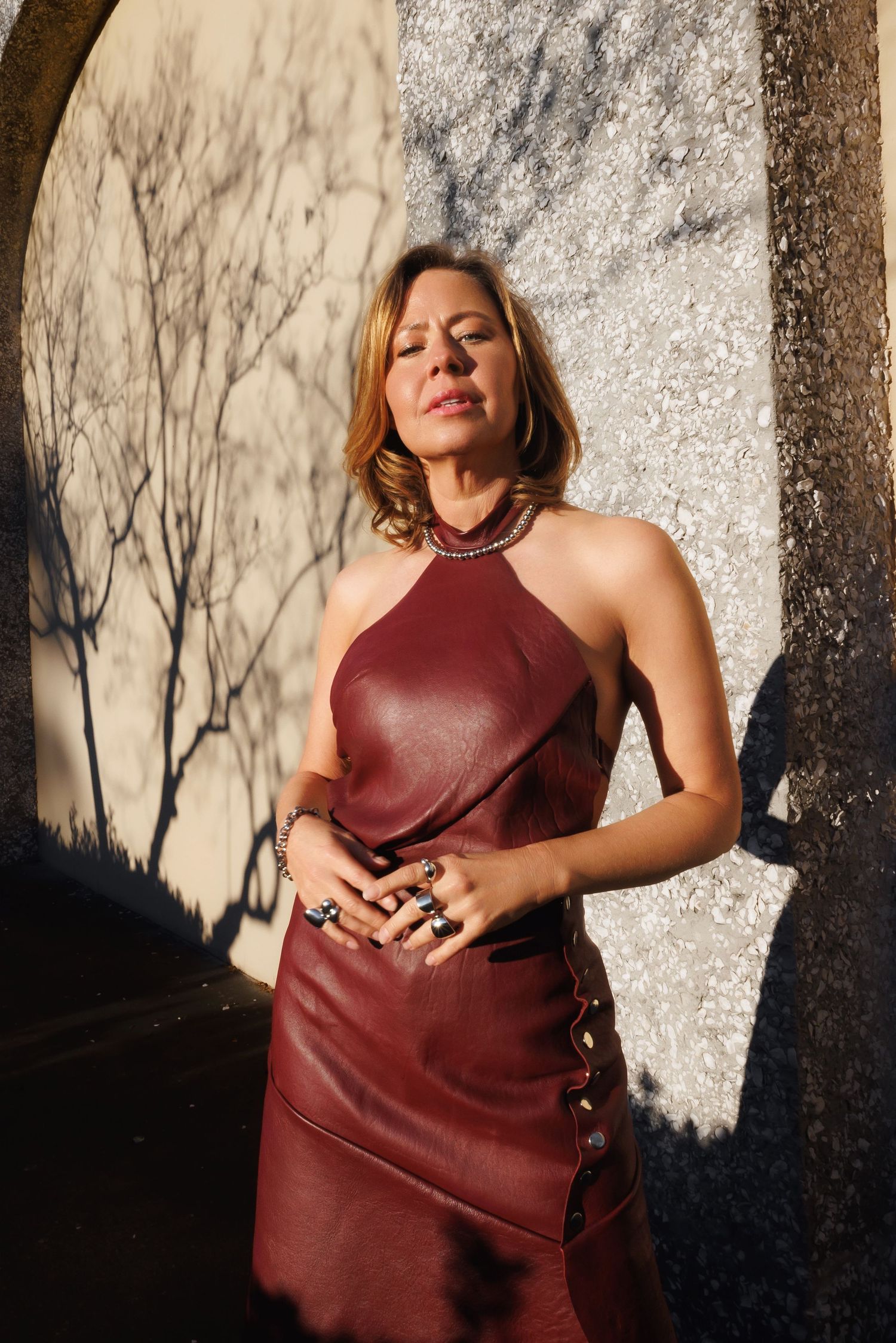 A dramatic fashion portrait series showing a model wearing a burgundy leather dress in dappled sunlight against a textured wall.