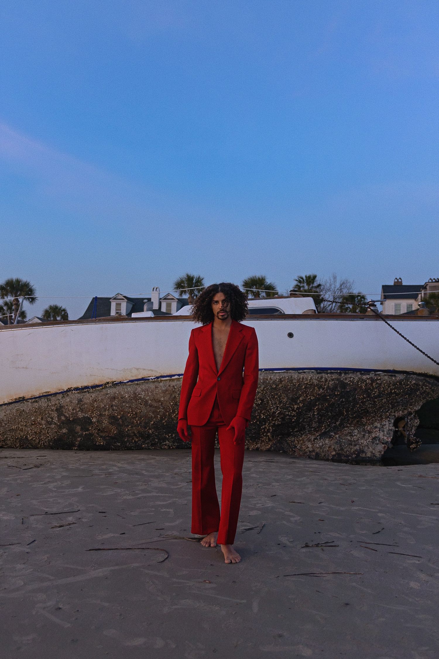 A figure in a striking red pantsuit stands barefoot on a beach at dusk with white buildings in the background.