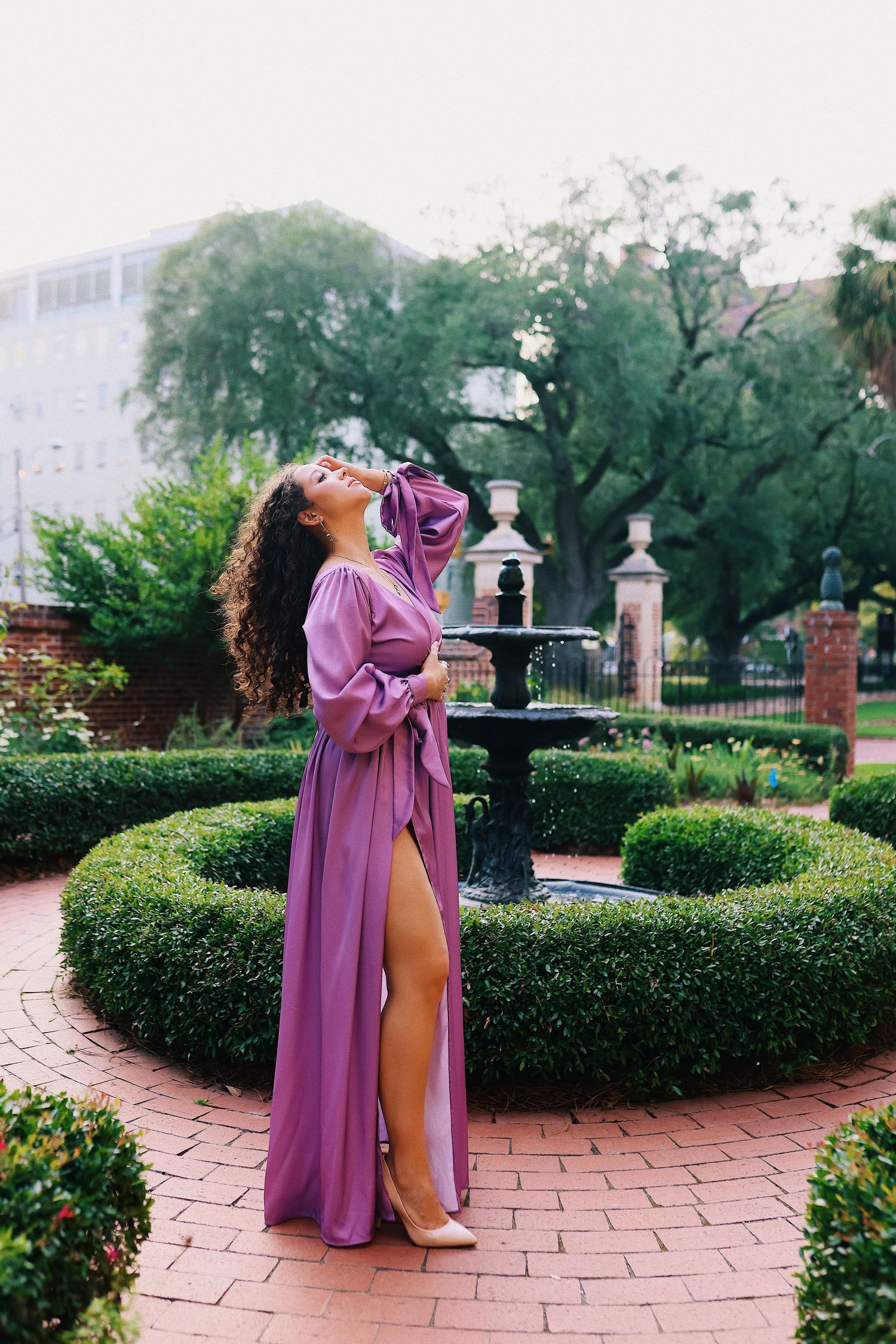 A person in a flowing pink robe stands in a formal garden with brick pathways and manicured hedges.