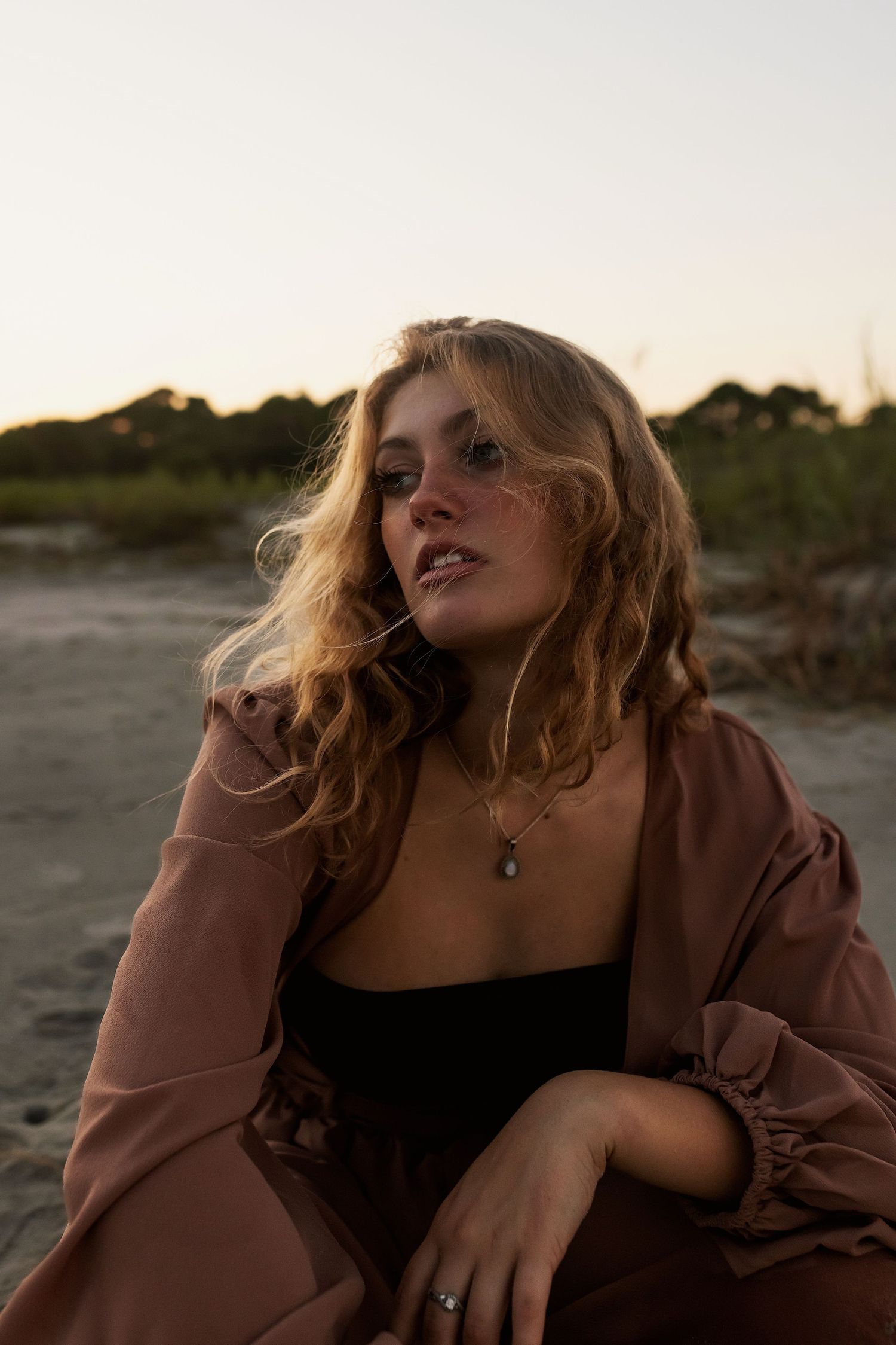 Atmospheric portrait at sunset with windswept hair wearing brown jacket and black top in natural outdoor setting.
