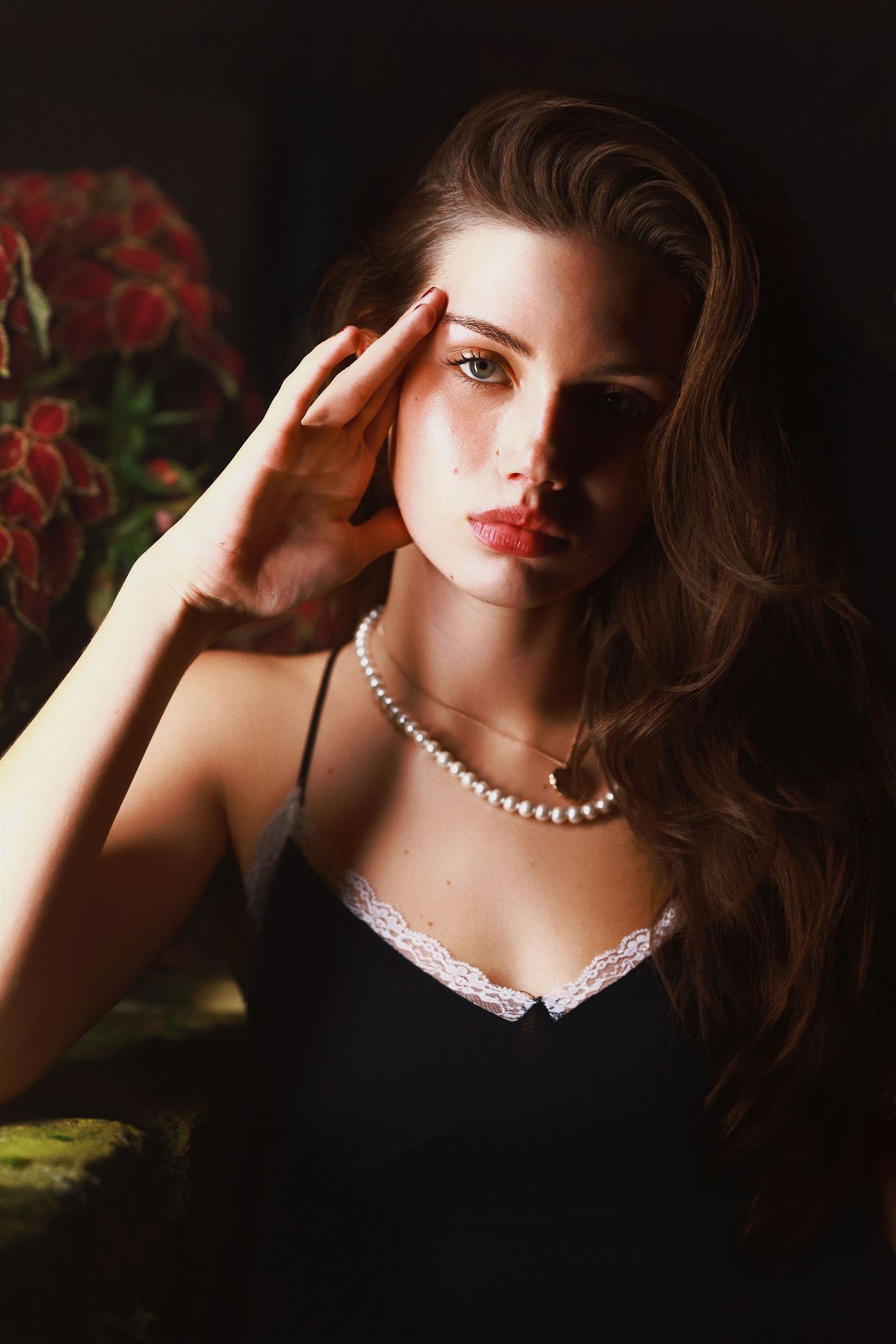 A glamorous portrait featuring pearls, red lipstick, and a black lace-trimmed dress in moody lighting with red flowers.
