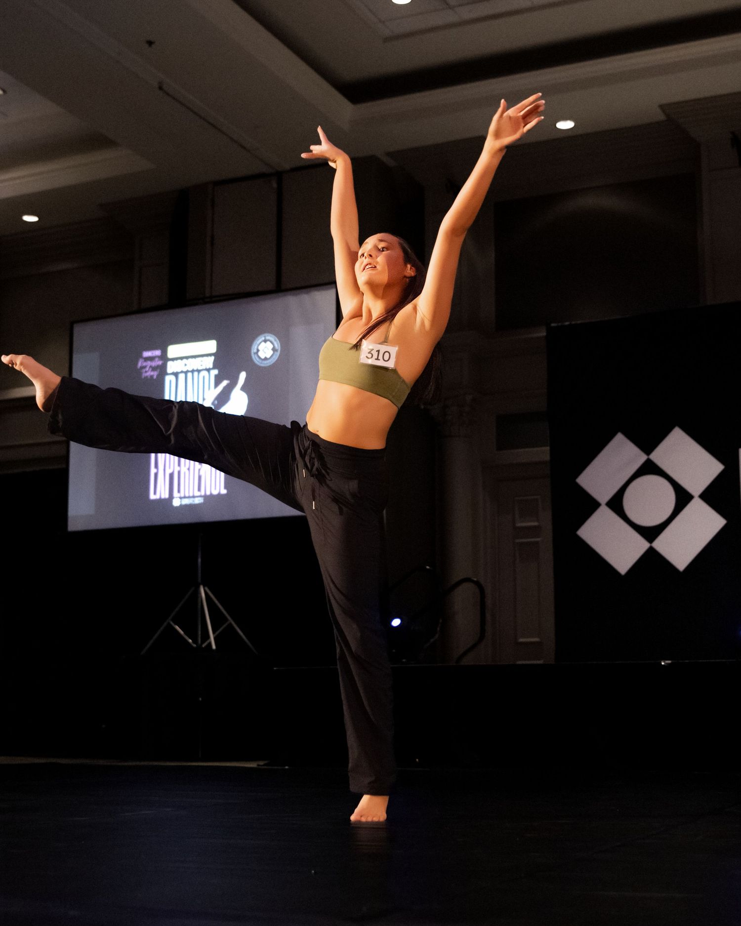A dancer performs an arabesque pose in a dark studio space with dramatic lighting and geometric shapes projected behind.