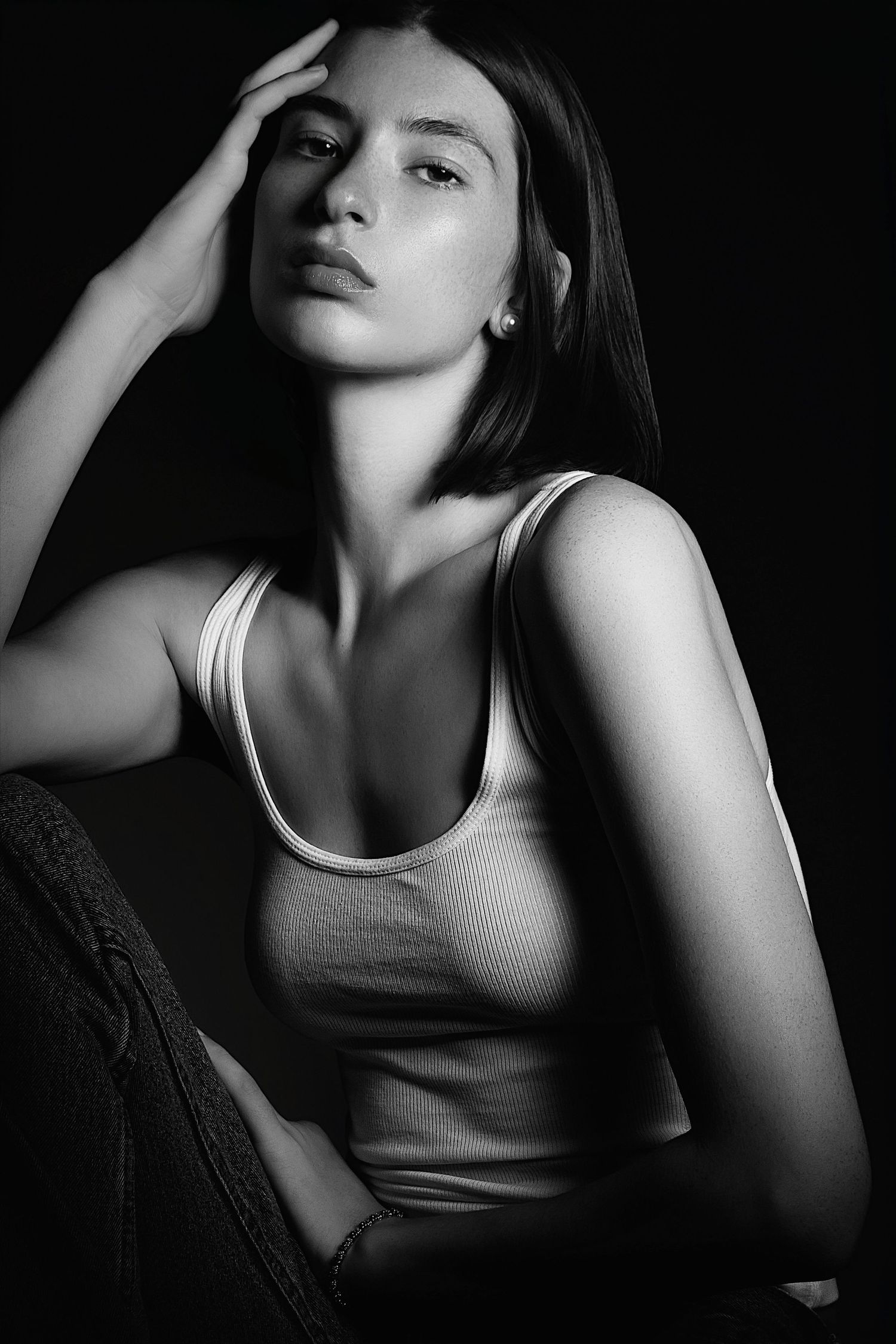 Black and white portrait shot with dramatic shadows showing a figure in a tank top with hand raised to their head.