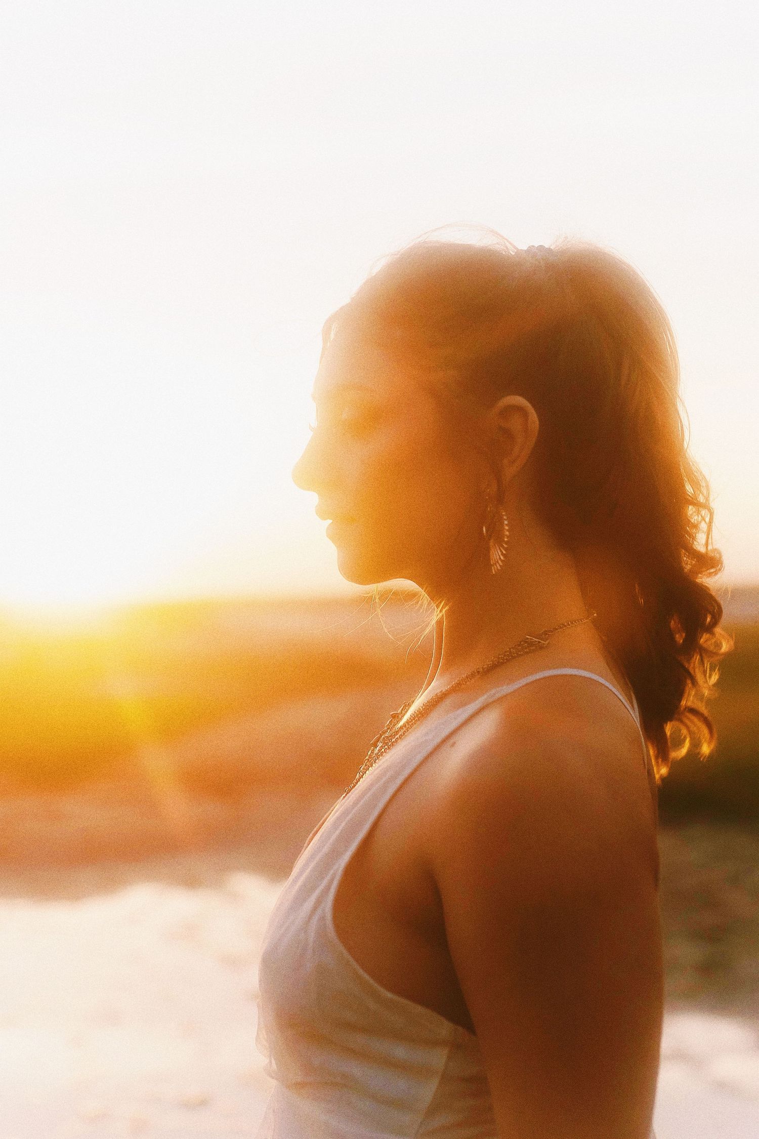 A silhouetted figure in a white tank top gazes at a warm sunset over water creating a dreamy golden atmosphere.