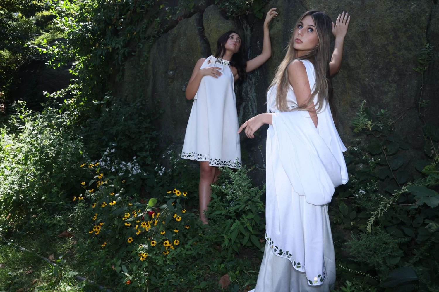 Two people in flowing white summer dresses pose playfully in a lush garden setting with yellow flowers.
