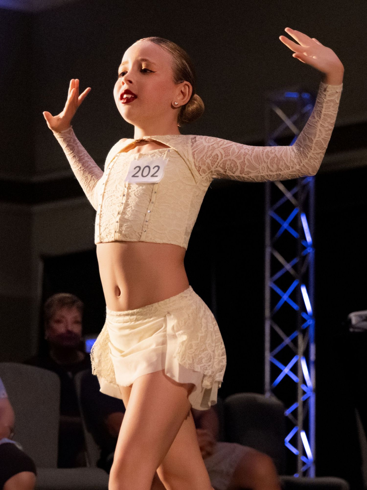 A dancer in white lace performs an expressive dance routine on stage with dramatic lighting in the background.