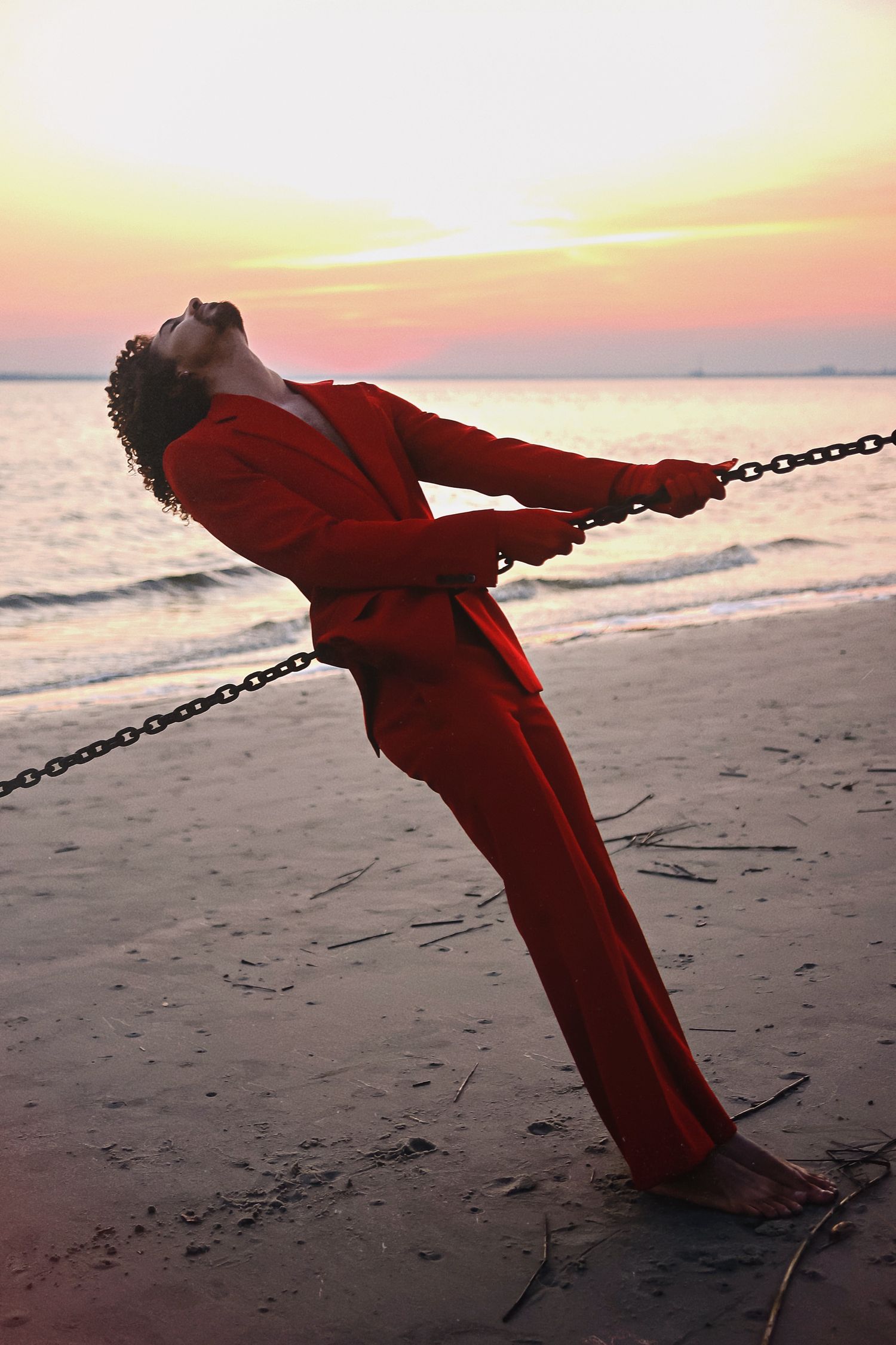 A silhouette in a red outfit leans back dramatically against a chain barrier on the beach at sunset.