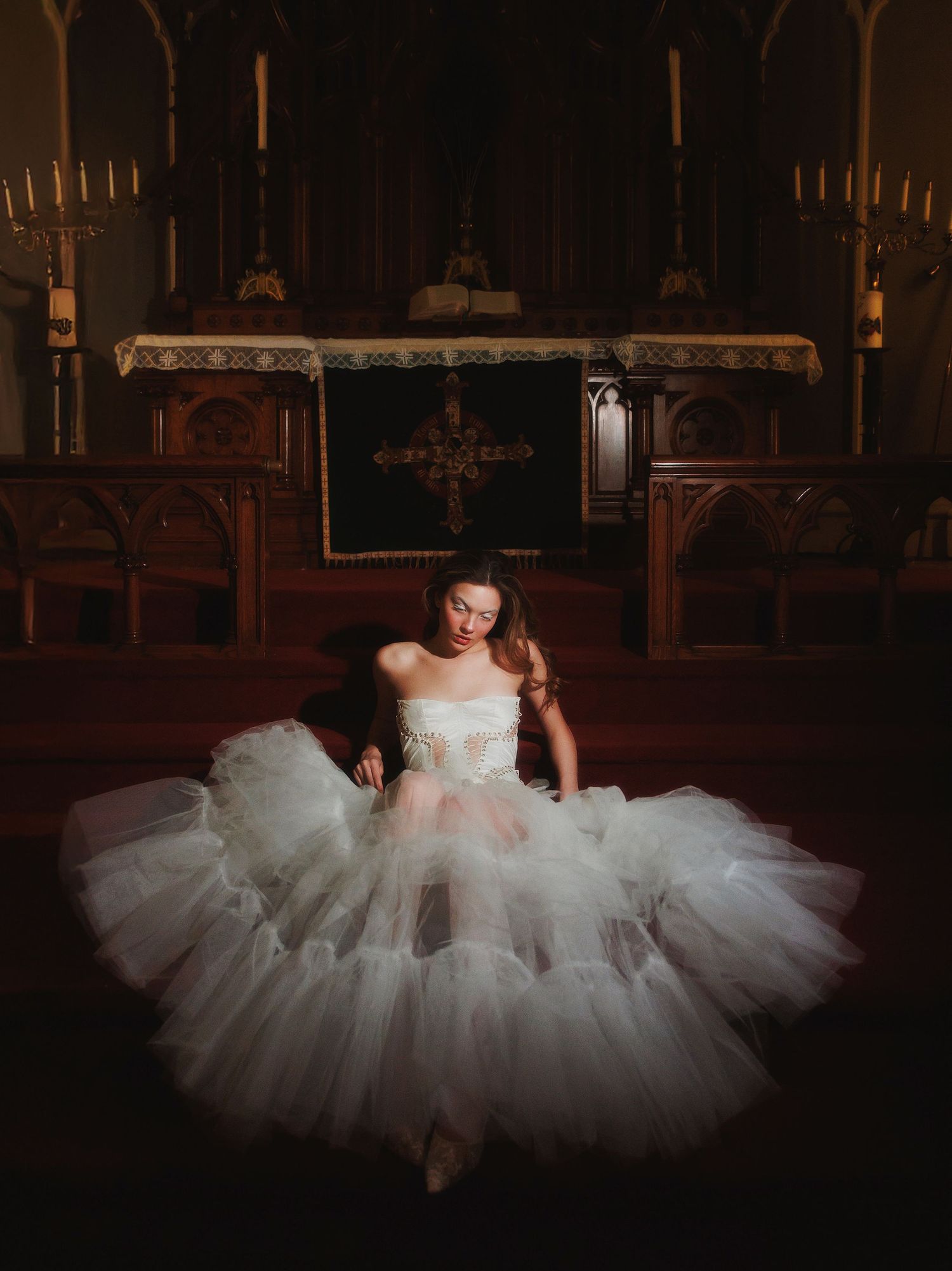 A white wedding dress with voluminous tulle skirt is photographed dramatically in a dark cathedral-like setting.